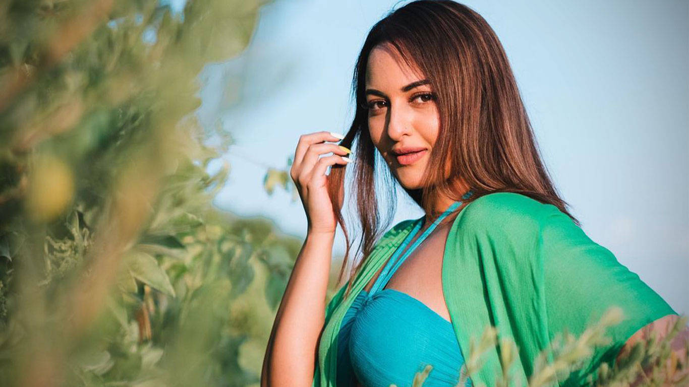 Sonakshi Sinha 綠色泳裝造型 壁紙 — 一位身穿鮮綠色和藍綠色服裝的女性，在明亮的日光下站在茂密的樹葉中，輕柔地觸摸頭髮，表情平靜。