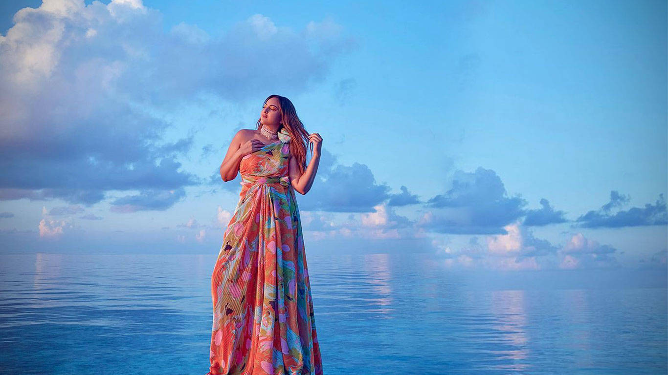 Sonakshi Sinha 長裙藍天 壁紙 — 一位身穿色彩繽紛飄逸長裙的女性，在日落時分，於柔和的藍天和散落的雲朵下，優雅地站在平靜的海面上。