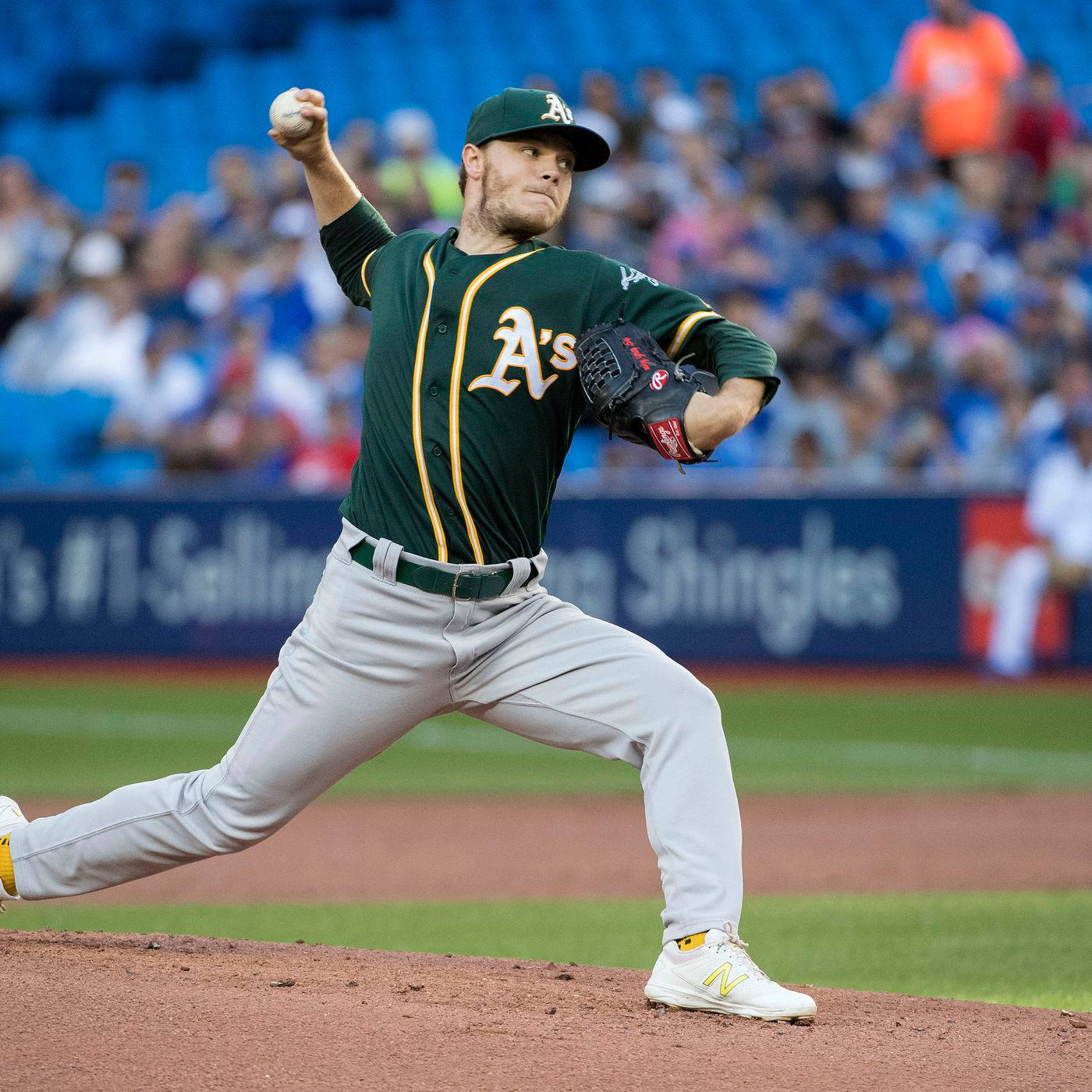 Sonny Gray 堅定投球 壁紙 — 一名身穿綠色和金色球衣的棒球投手，在陽光普照的球場上投球，背景有藍色座位上的模糊觀眾。