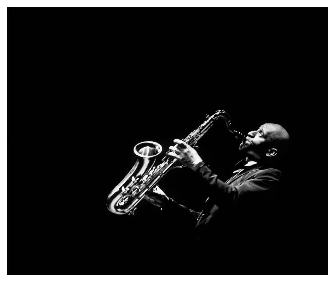 傳奇爵士薩克斯風手 Sonny Rollins 演奏時的畫面 壁紙 — 一位爵士音樂家在黑色背景的戲劇性舞台燈光下熱情地演奏色士風。