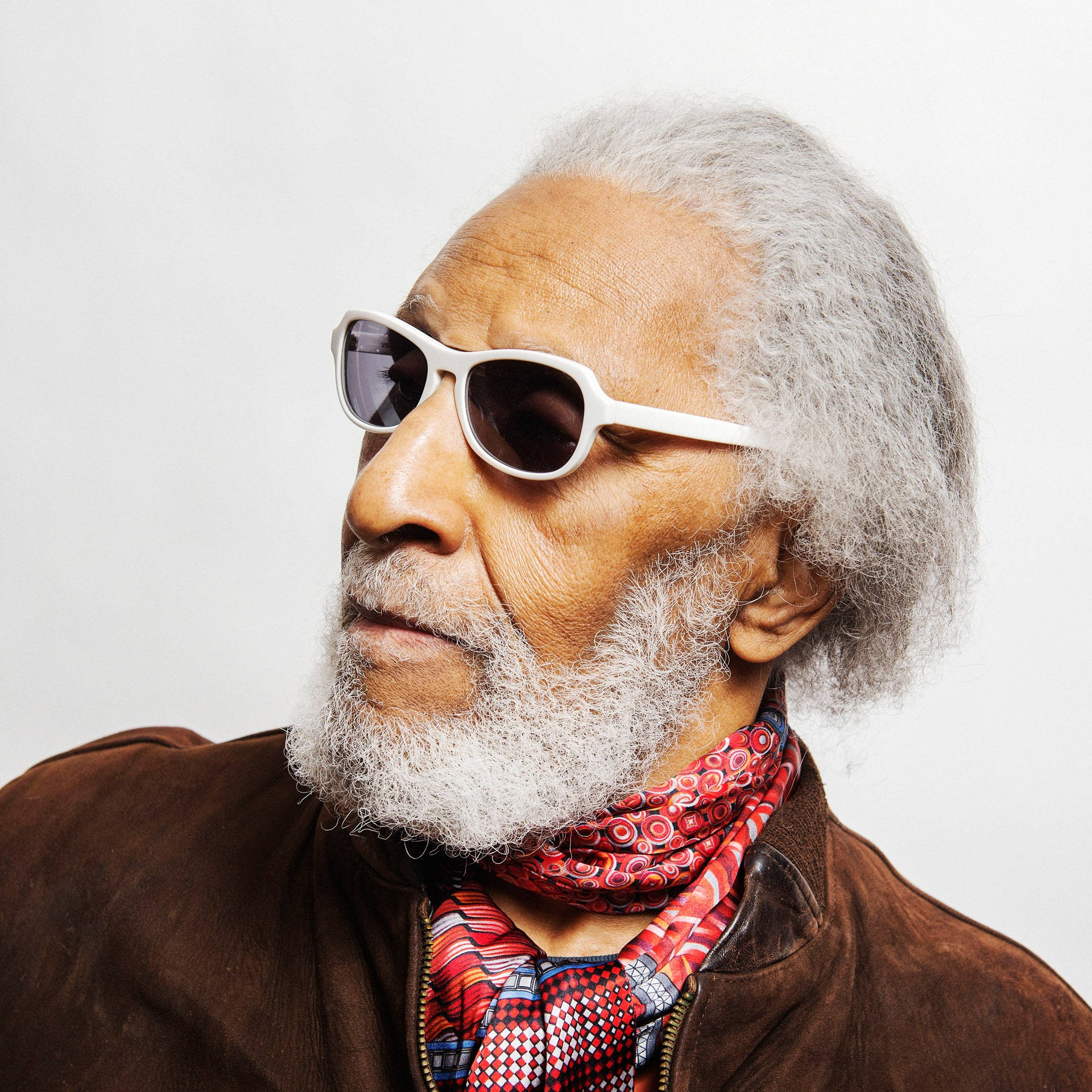 Sonny Rollins 的近距離肖像 壁紙 — 一位時尚的老年男性，白髮白鬍，戴著白色太陽眼鏡和彩色圖案圍巾，背景為素色。