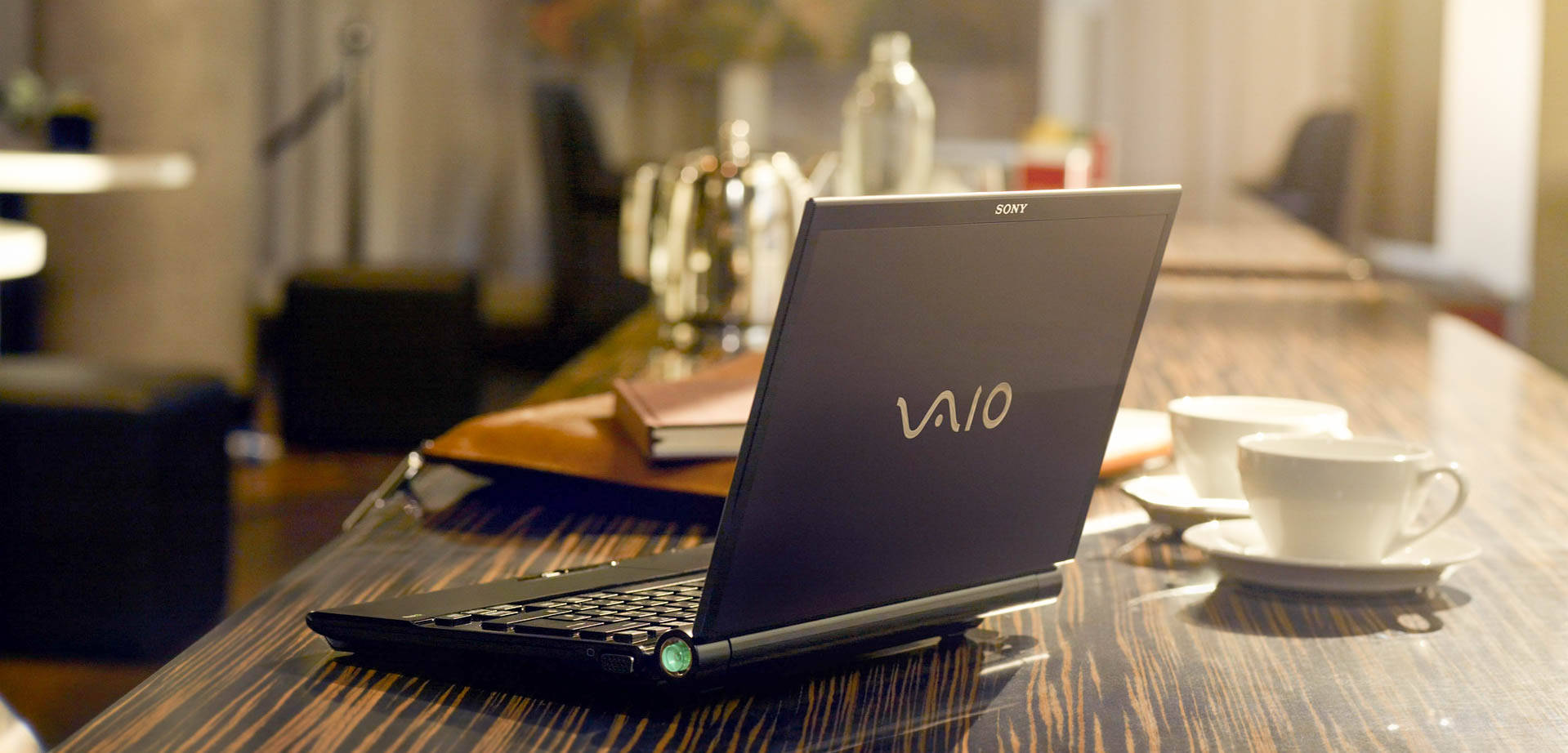Sony產品：VAIO筆記本電腦與辦公桌 壁紙 — 一部時尚的黑色Sony Vaio筆記型電腦打開放在深色木桌上，旁邊有兩個白色咖啡杯，營造出舒適的咖啡館氛圍。