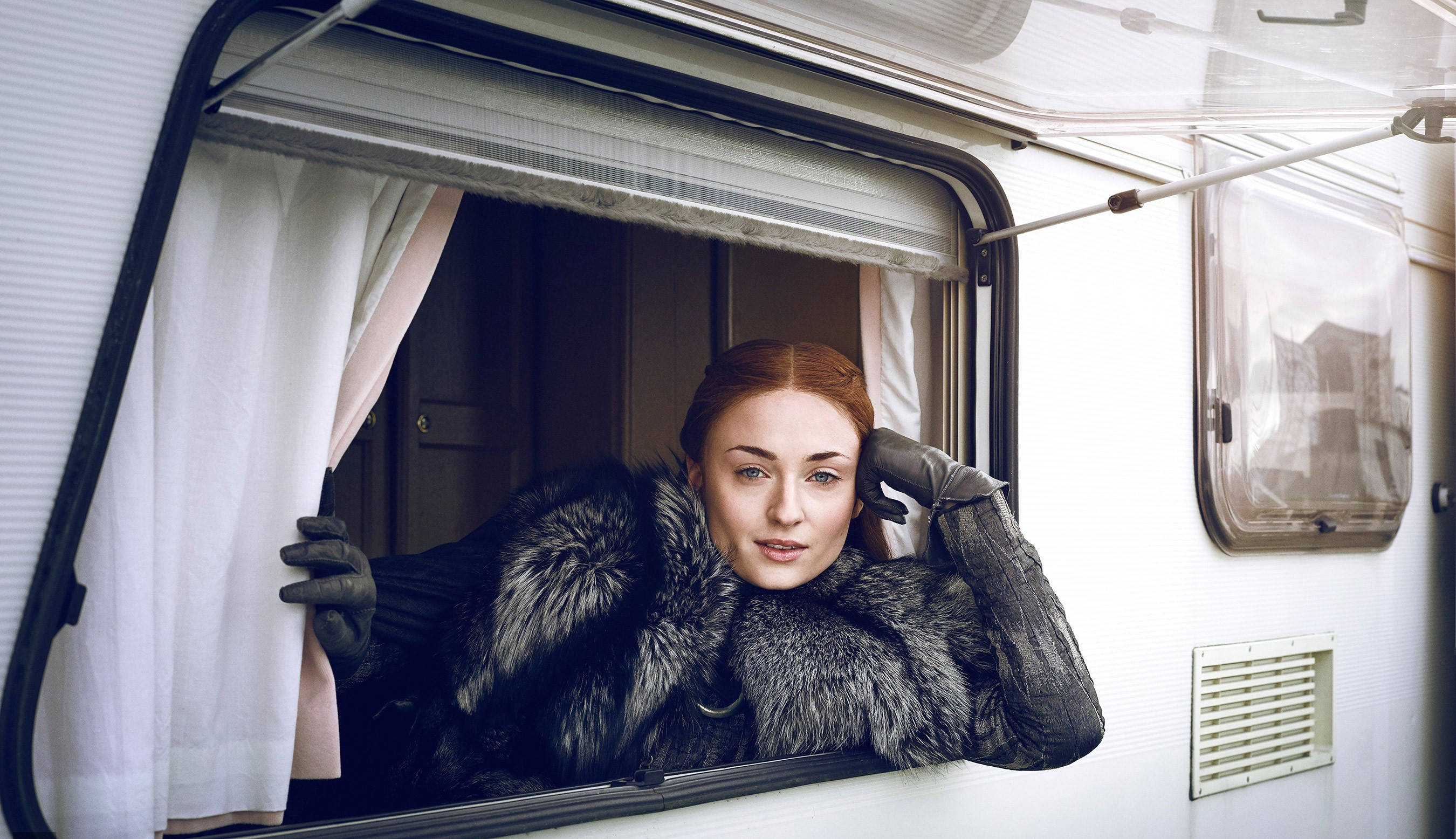 Sophie Turner 飾演 Sansa Stark 壁紙 — 一位身穿毛領大衣的女子，從露營車窗探出身子，手持窗簾，神情平靜。