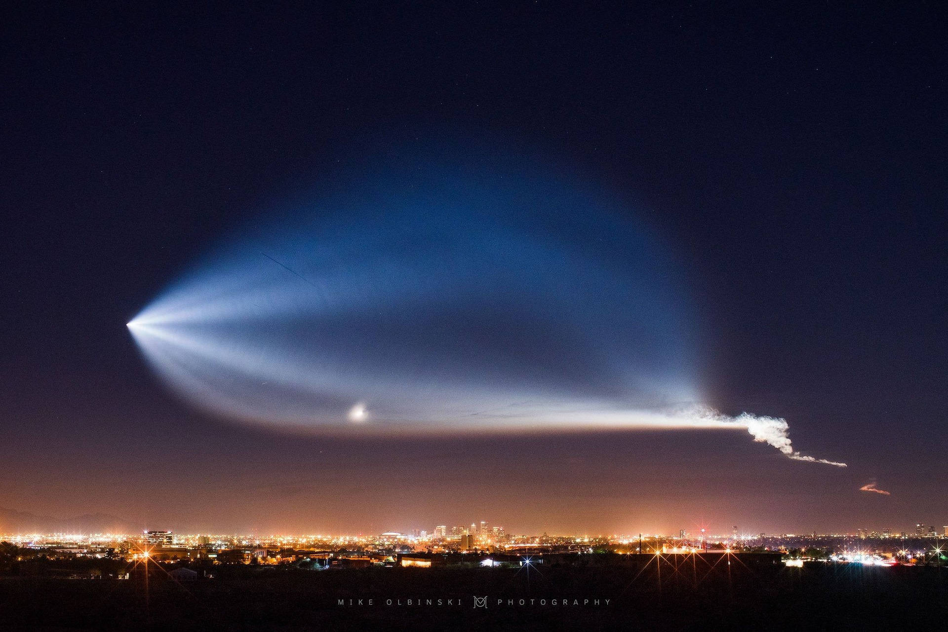 一條明亮的藍色凝結尾跡照亮城市上空的夜空。 — Spacex GIF 壁紙