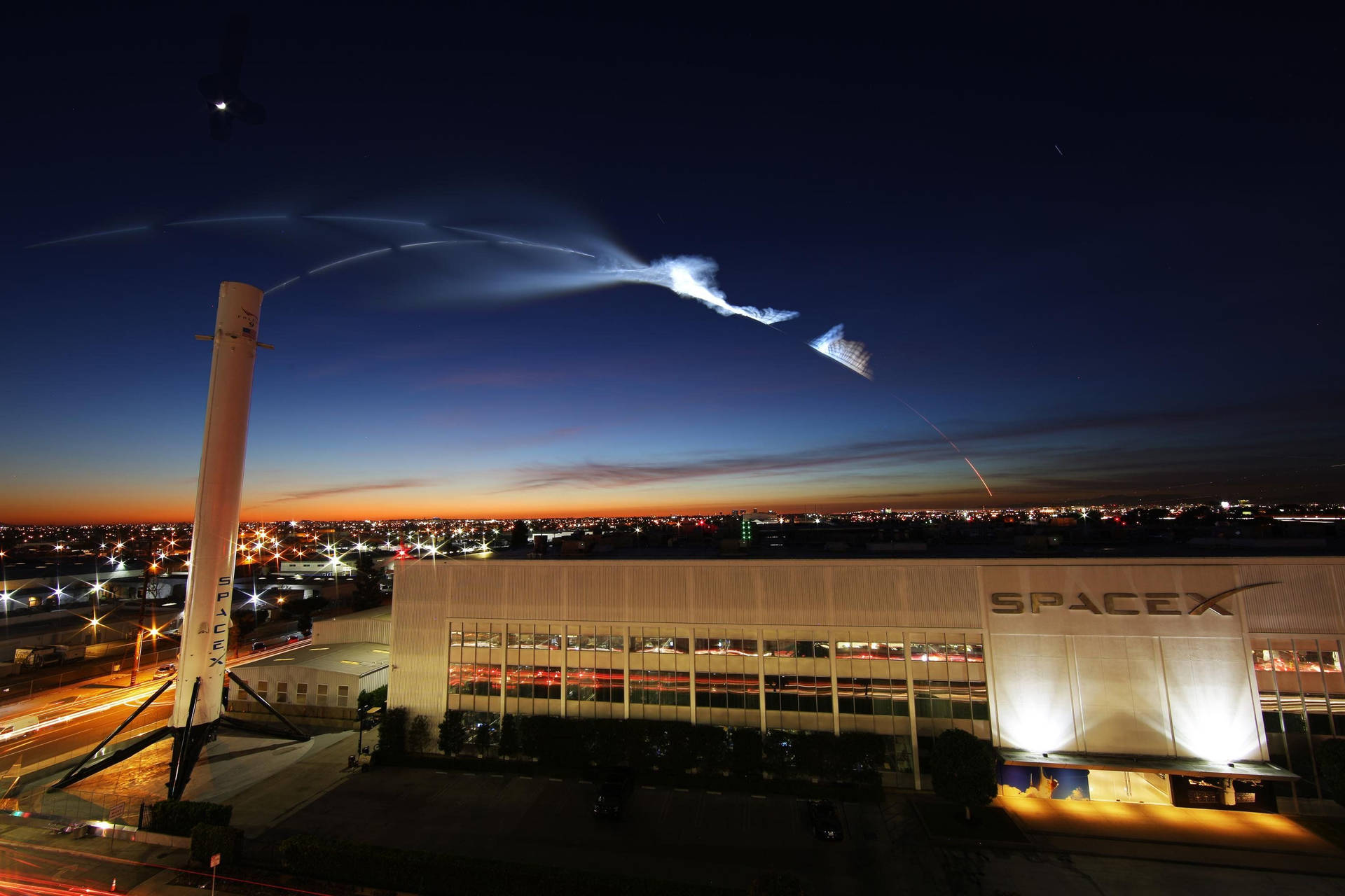 SpaceX 火箭在黃昏時於燈火通明的設施上空發射，下方有城市燈光。 — Spacex GIF 壁紙