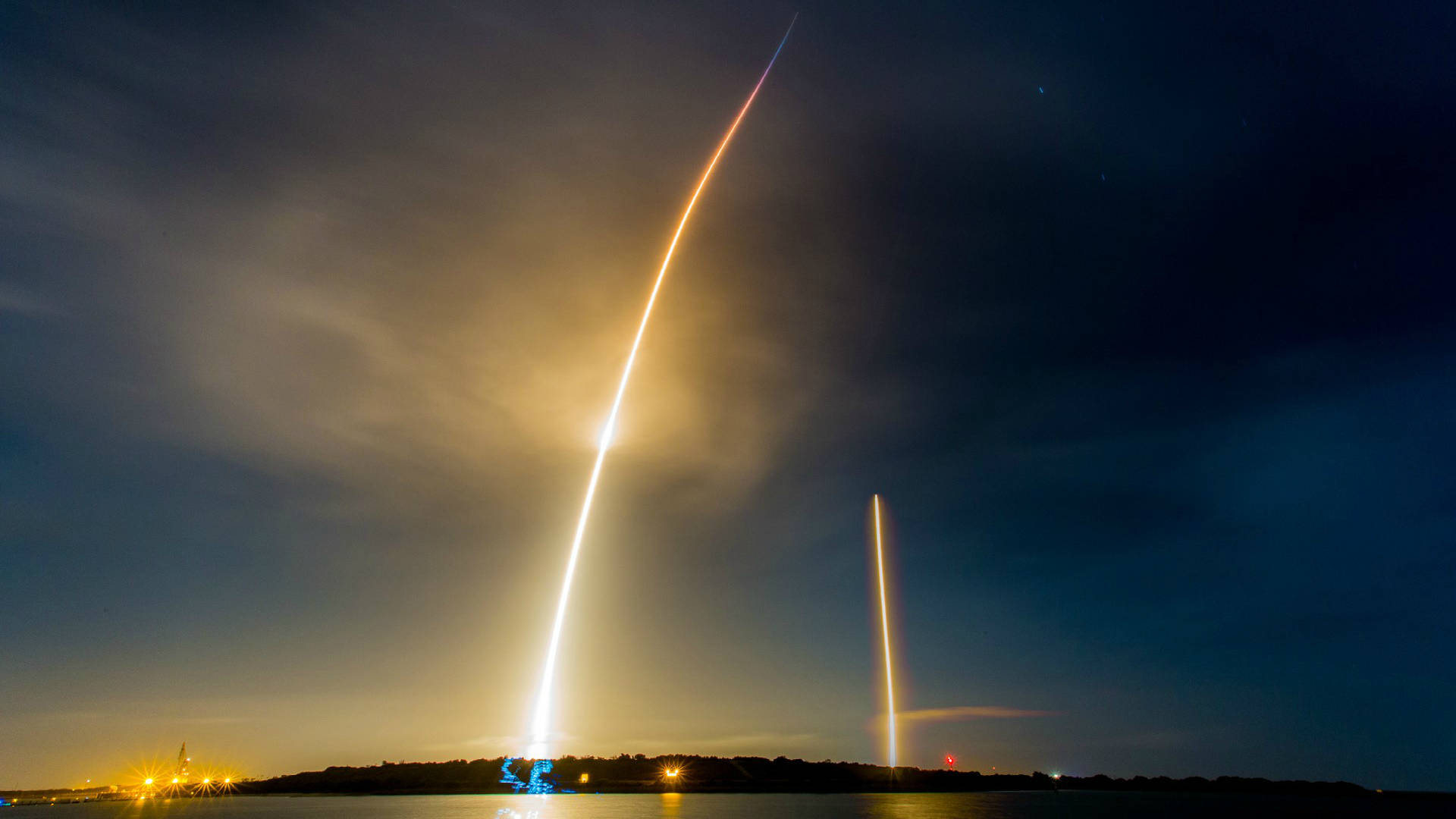 夜間火箭發射，水面上有明亮的光跡和遠處的燈光。 — Spacex GIF 壁紙