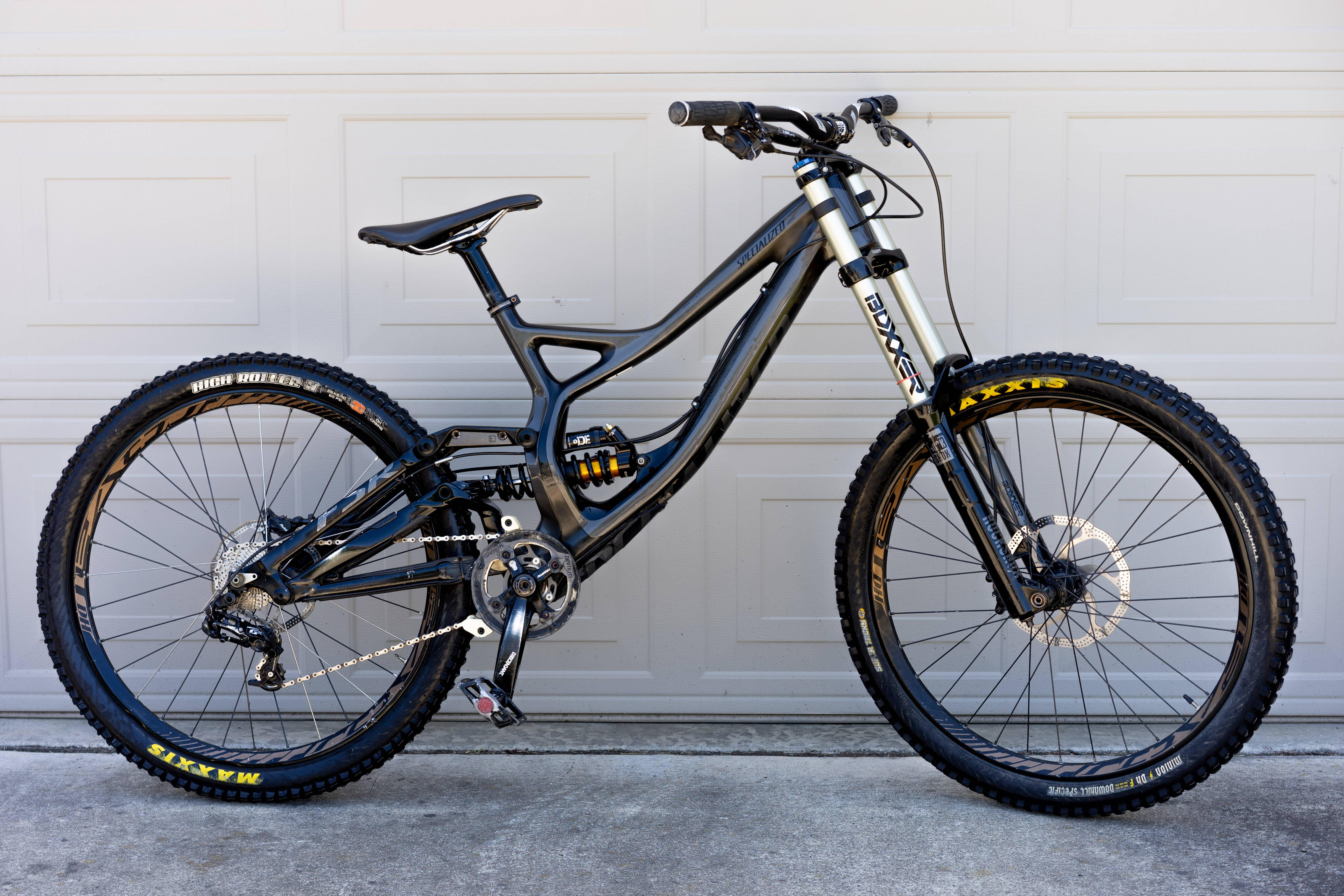 Specialized 黑色山地自行車 壁紙 — 一架時尚的黑色全避震山地單車，配備黃色Maxxis輪胎，停放在白色車庫門前。