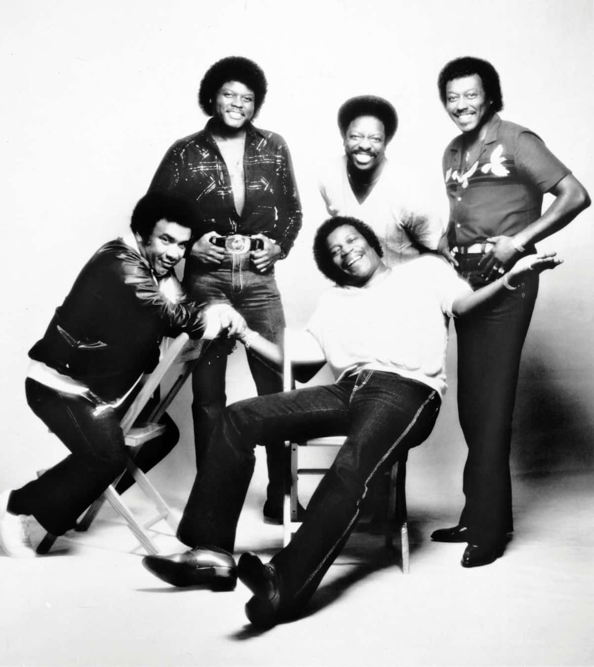 Spinners 的單色室內照片 壁紙 — 五位微笑男士身穿1970年代時尚，在簡約的攝影棚背景前與椅子擺出俏皮姿勢。