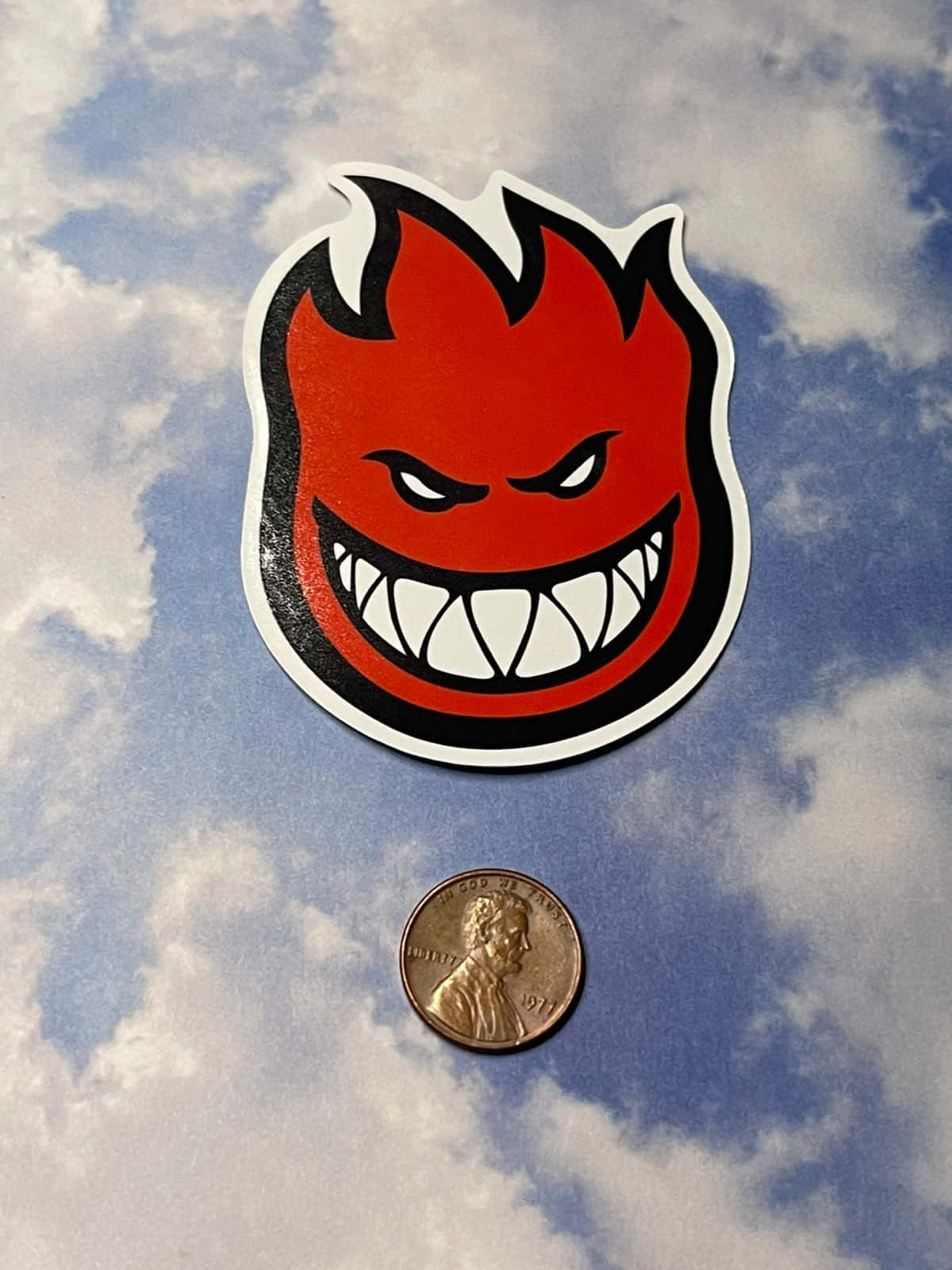 紅色魔鬼貼紙，有尖銳牙齒，位於美國一美分硬幣上方，背景為多雲天空。
