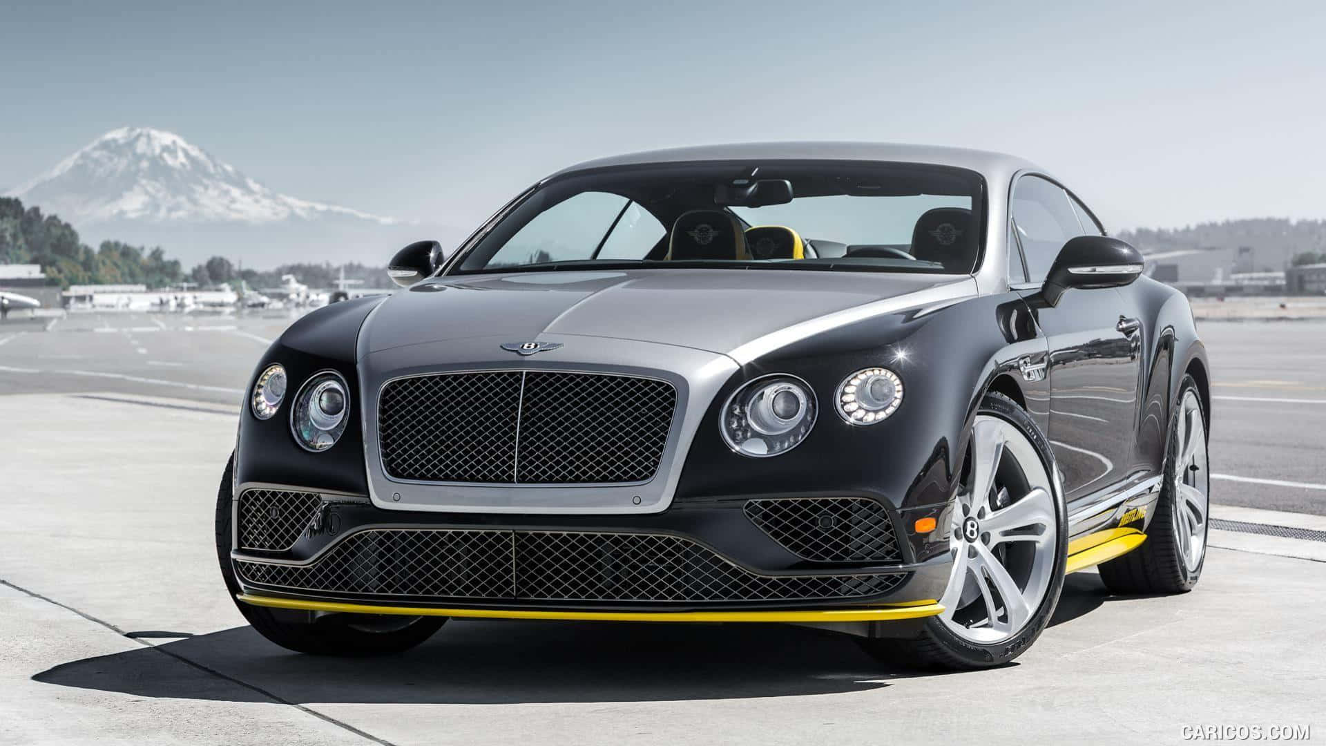 賓利Sport Breitling Edition 壁紙 — 一架時尚的黑色 Bentley Continental GT 帶有黃色點綴，停在機場停機坪上，背景是白雪覆蓋的山峰。