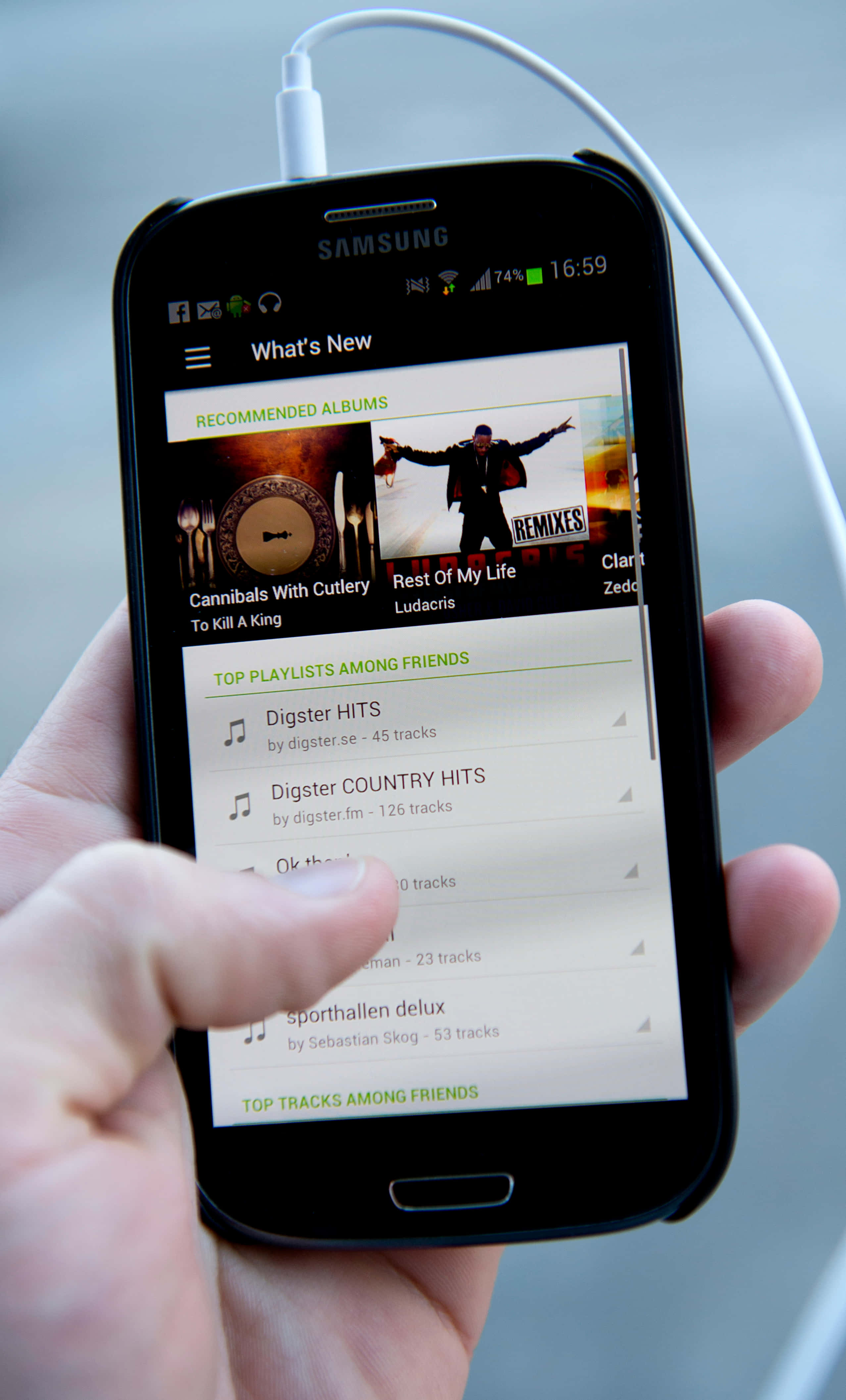 Spotify 新歌推薦截圖 壁紙 — 一隻手拿著一部黑色Samsung智能手機，顯示一個音樂應用程式，包含播放清單和專輯封面，同時有一條白色充電線連接到頂部。