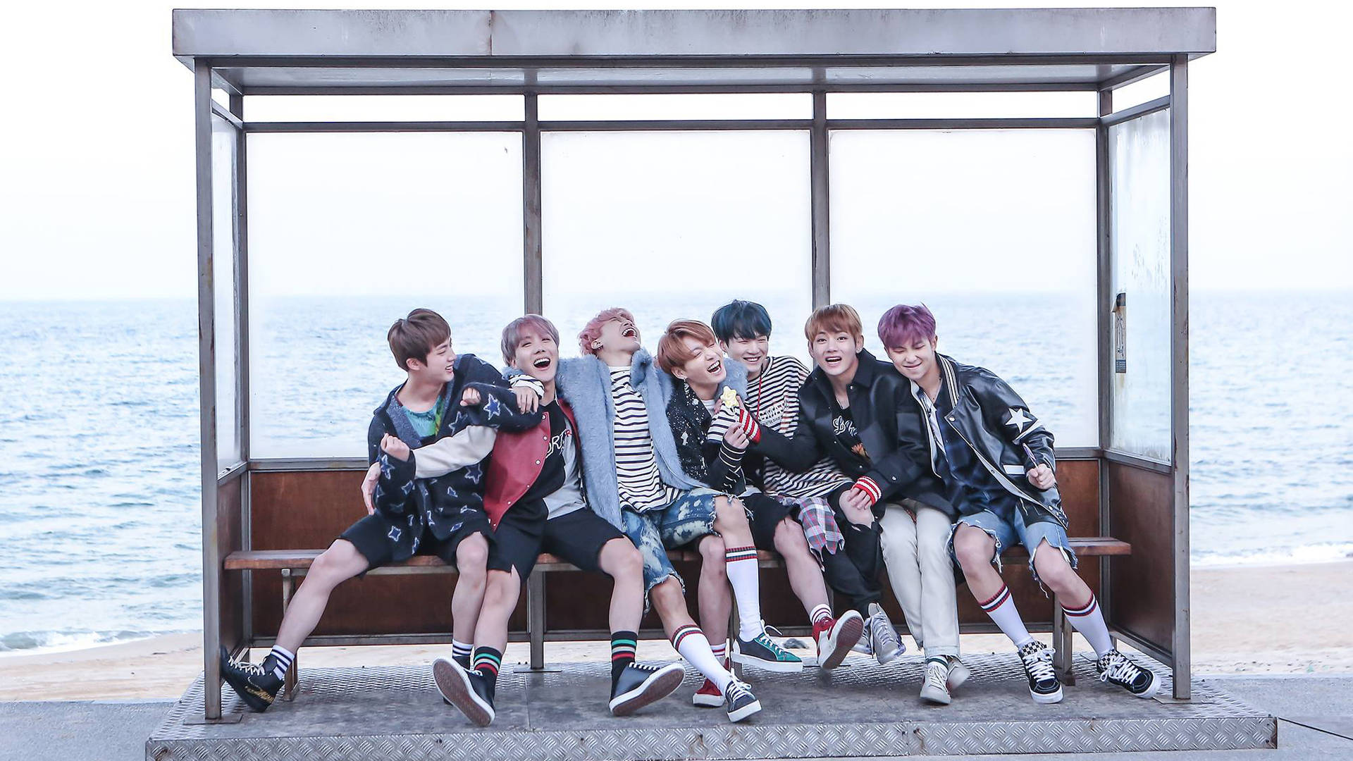 《Spring Day》預告酷炫BTS筆電壁紙 壁紙 — 七位身穿時尚街頭服的年輕男士在海邊巴士站長椅上大笑並擺姿勢，身後是海洋。