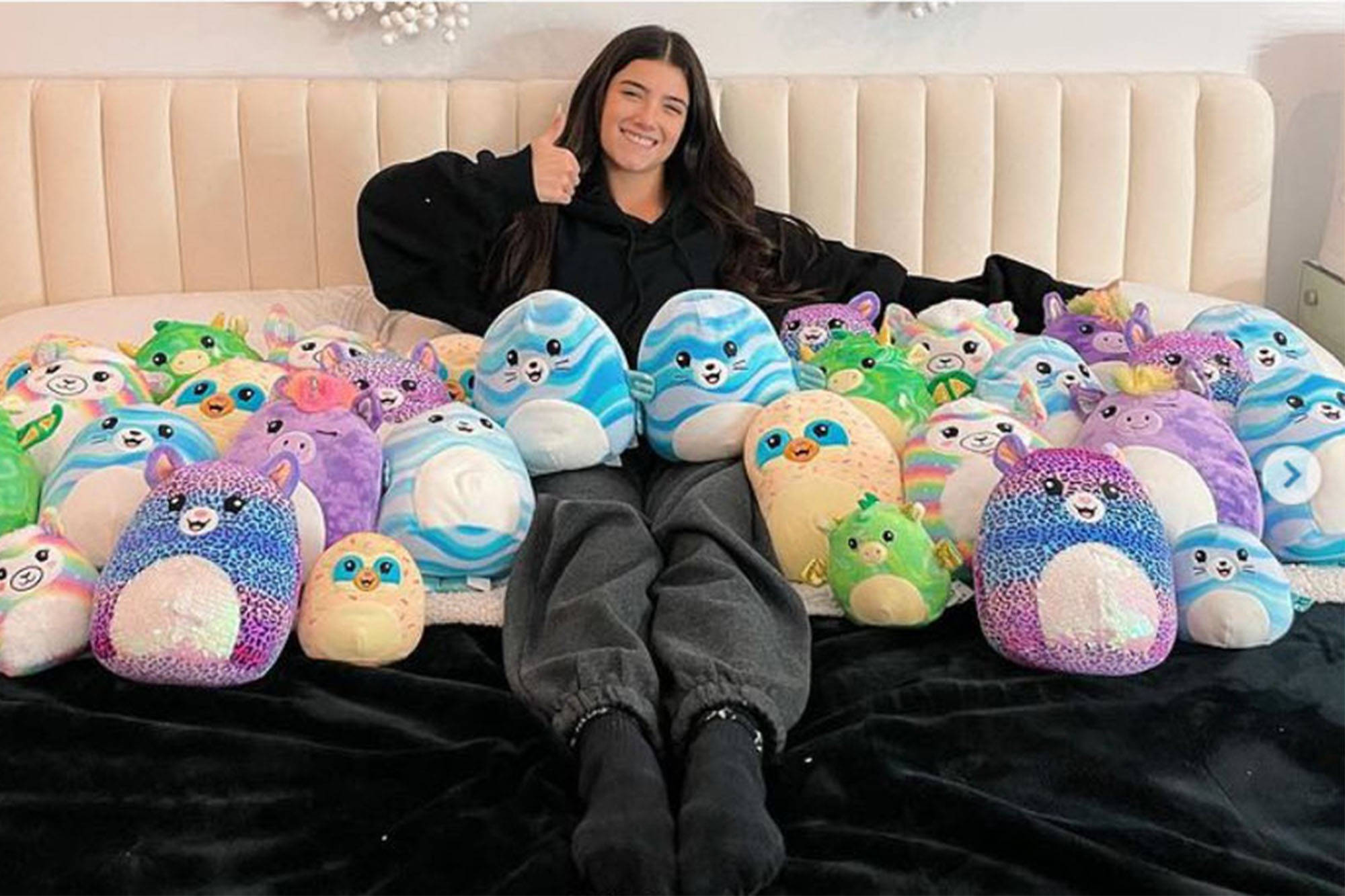 與 Squishmallows 玩具共處的女性 壁紙 — 一位微笑的年輕女性坐在床上，豎起大拇指，周圍是色彩繽紛的軟膠玩具收藏。
