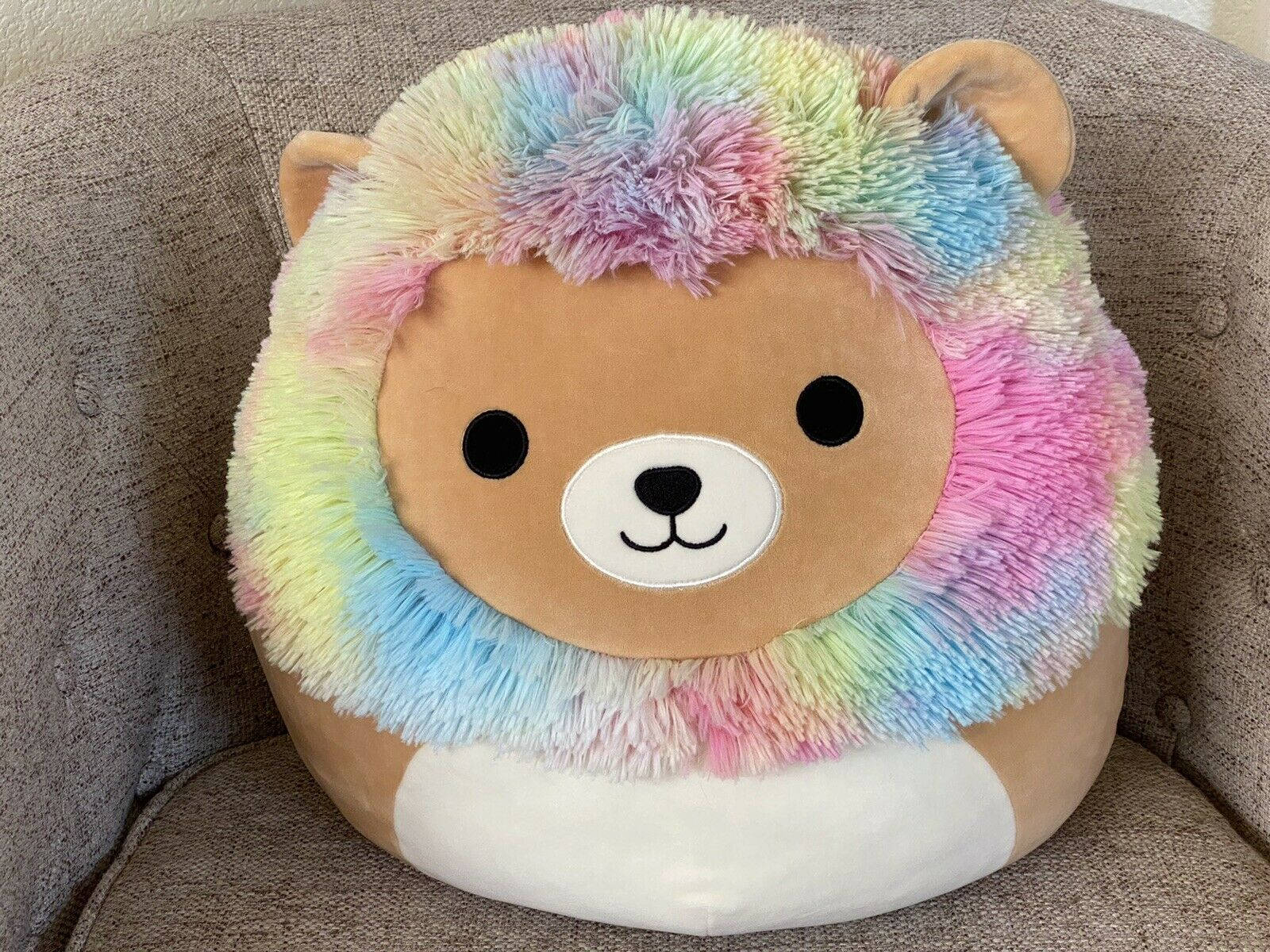Squishmallows獅子在沙發上 壁紙 — 一隻彩虹毛絨獅子幼崽舒適地坐在有紋理的灰色沙發墊上。