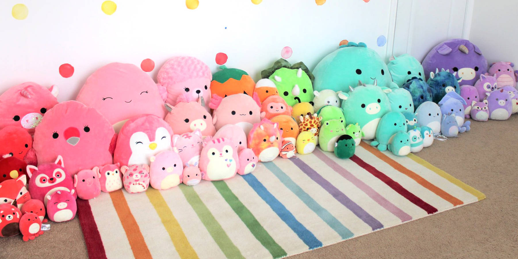 Squishmallows玩偶收藏 壁紙 — 一系列色彩繽紛的柔軟 squishmallows 以彩虹條紋排列，背景為白色圓點牆。