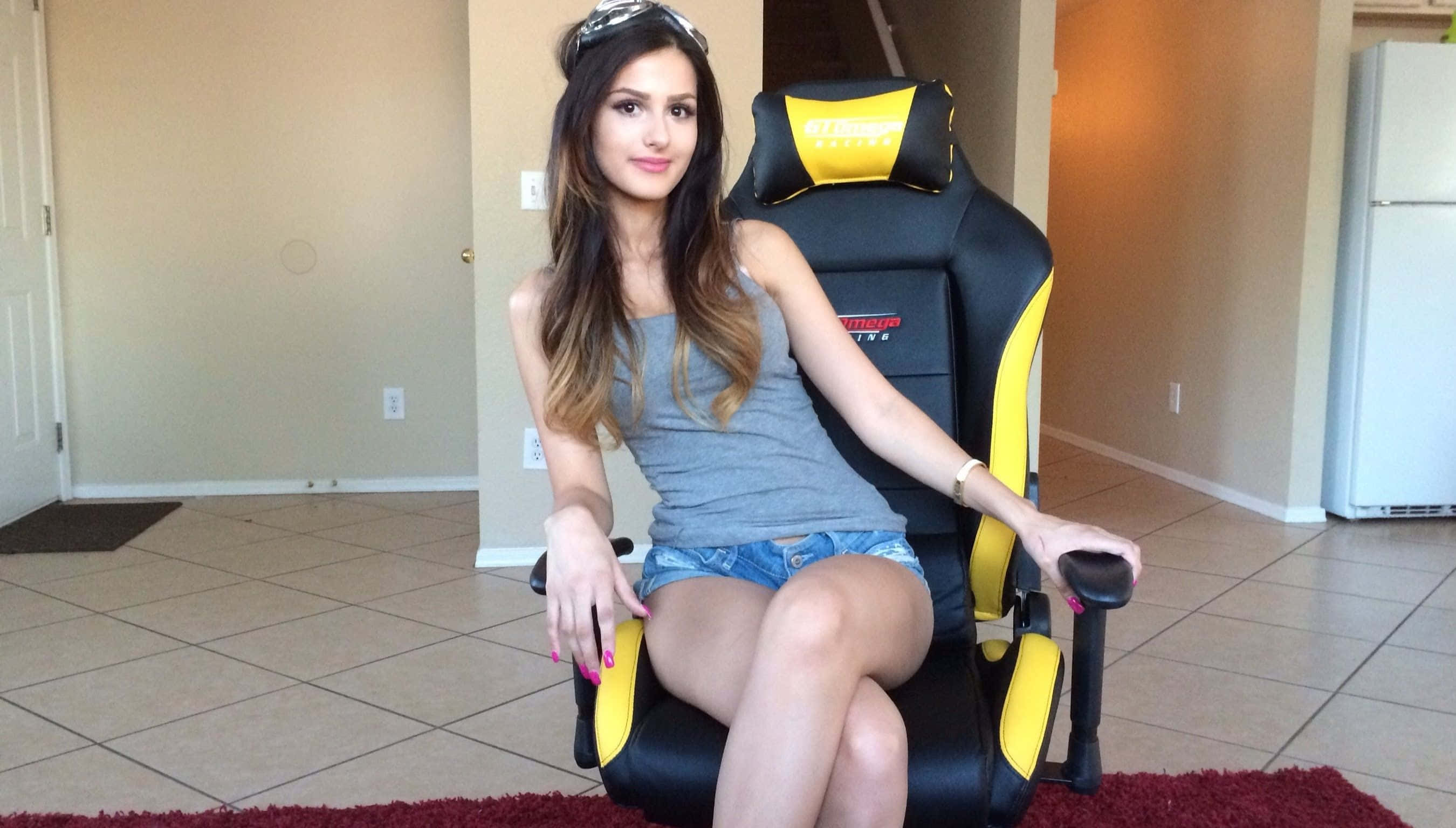 Sssniperwolf 遊戲椅 壁紙 — 一位長髮年輕女性自信地坐在舖有瓷磚的房間裡的黑色和黃色電競椅上。