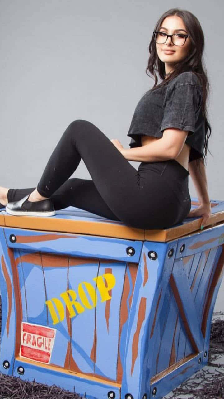 Sssniperwolf 藍色貨箱手機殼 壁紙 — 一位戴眼鏡的女性隨意地坐在一個藍色油漆的運輸箱上，箱子上標有 'DROP' 和 'FRAGILE'，背景是純灰色。