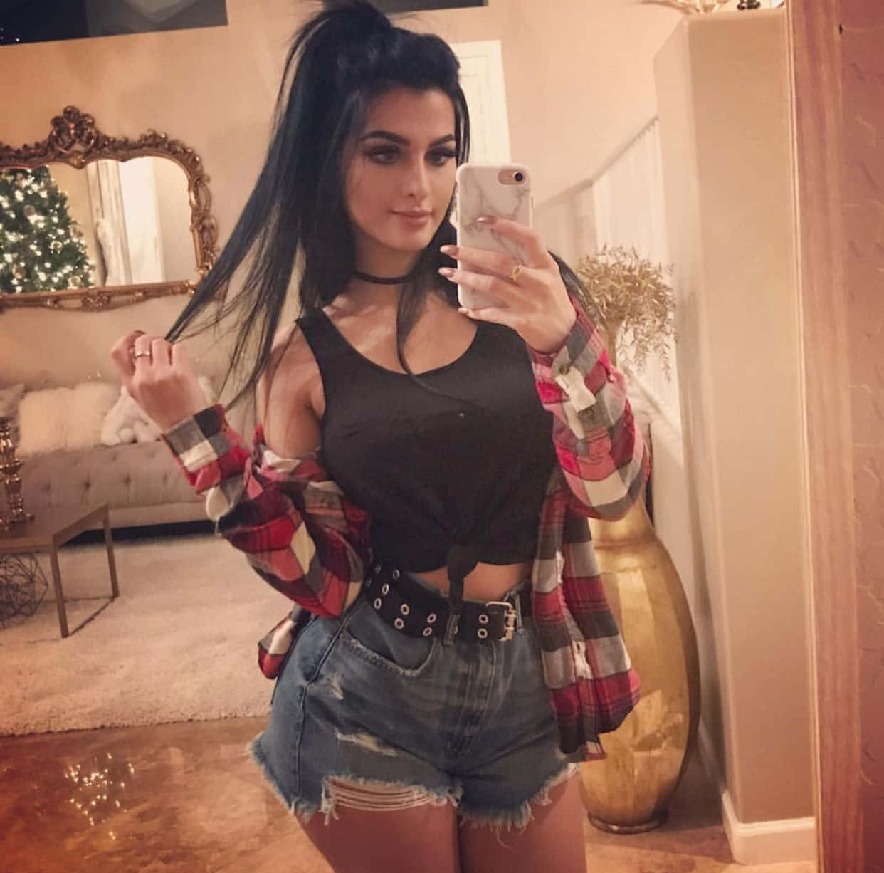 Sssniperwolf 鏡前自拍 壁紙 — 一位深色頭髮的女性在溫暖照明的客廳中，穿著格仔襯衫和牛仔短褲，對著鏡子自拍。