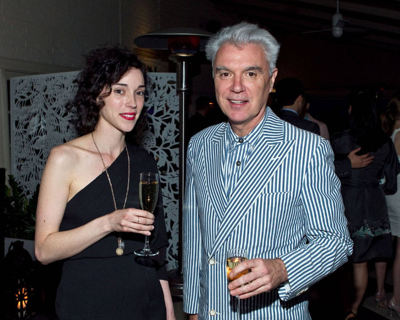 大衛·拜恩與 St. Vincent（艾琳·克拉克） 壁紙 — 一男一女在光線昏暗的室內派對中微笑，手持香檳杯，周圍有裝飾屏風和溫暖的燈光。