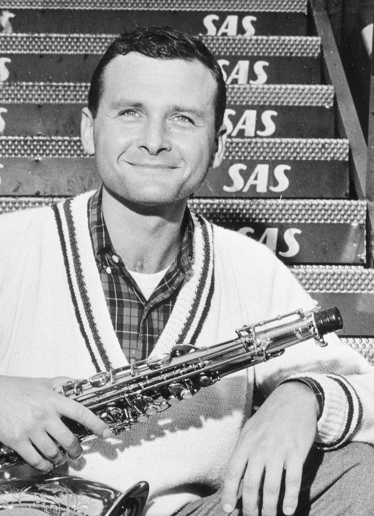 Stan Getz 在哥本哈根卡斯特魯普機場 壁紙 — 一個微笑的男人穿著毛衣，在樓梯上重複的SAS標誌背景下拿著薩克斯風。