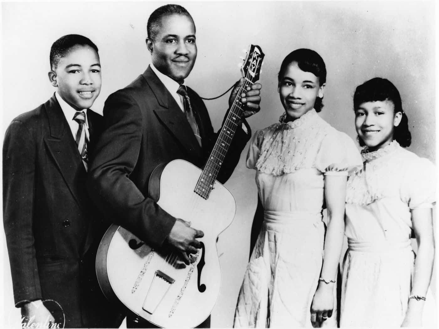 美國 Staple Singers 1955 年攝影棚肖像 壁紙 — 一個復古黑白工作室肖像，一個四口之家面帶微笑，穿著正式，其中一名男子手持原聲結他。