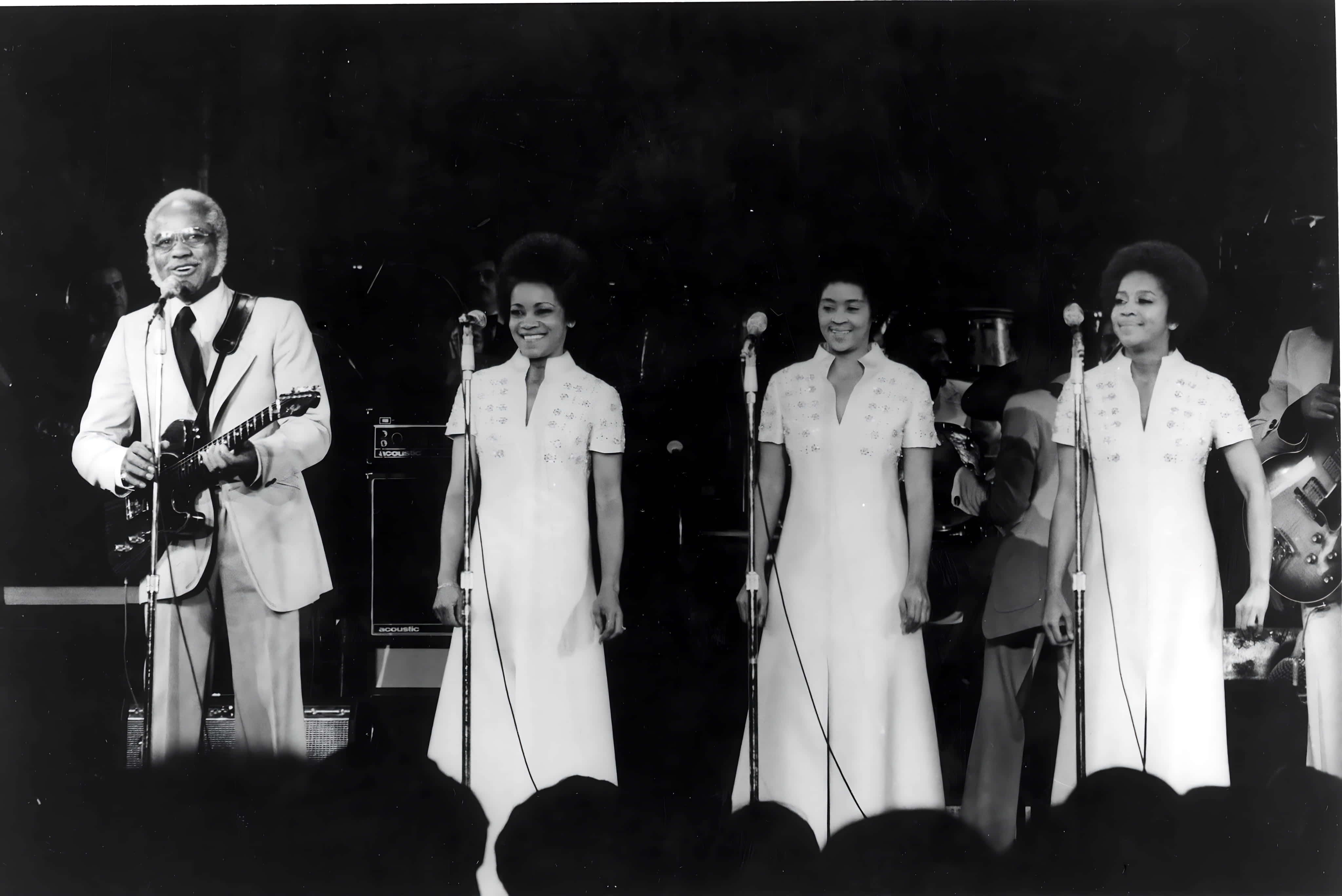 美國 Staple Singers 1970 年演唱會現場照片 壁紙 — 一張黑白照片，一名男結他手和三名身穿白色連衣裙的女歌手在舞台上用咪高峰表演。
