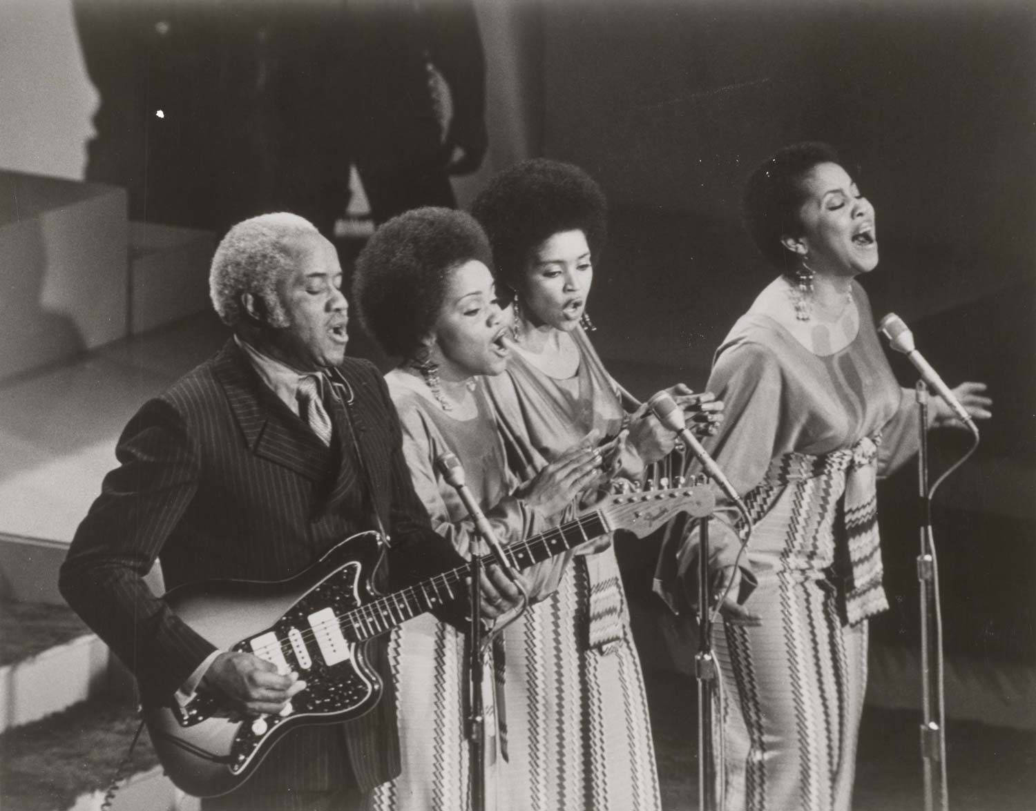 美國組合「 Staple Singers 」1971年《音樂改變一切的年份》現場演出 壁紙 — 一張黑白照片，拍攝一位男結他手和三位女歌手在舞台上手持麥克風熱情演出。