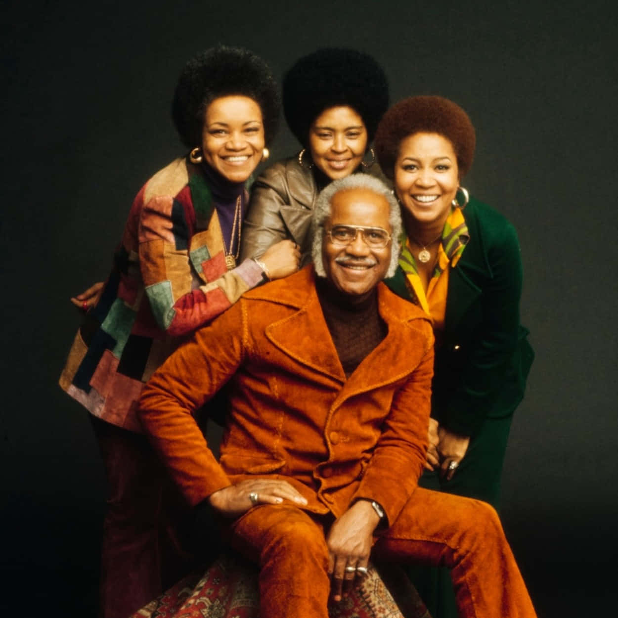 Staple Singers《1986》專輯封面 壁紙 — 四位面帶微笑、留著非洲式髮型的人，穿著鮮豔的70年代服裝，在深色攝影棚背景下溫馨地擺姿勢。