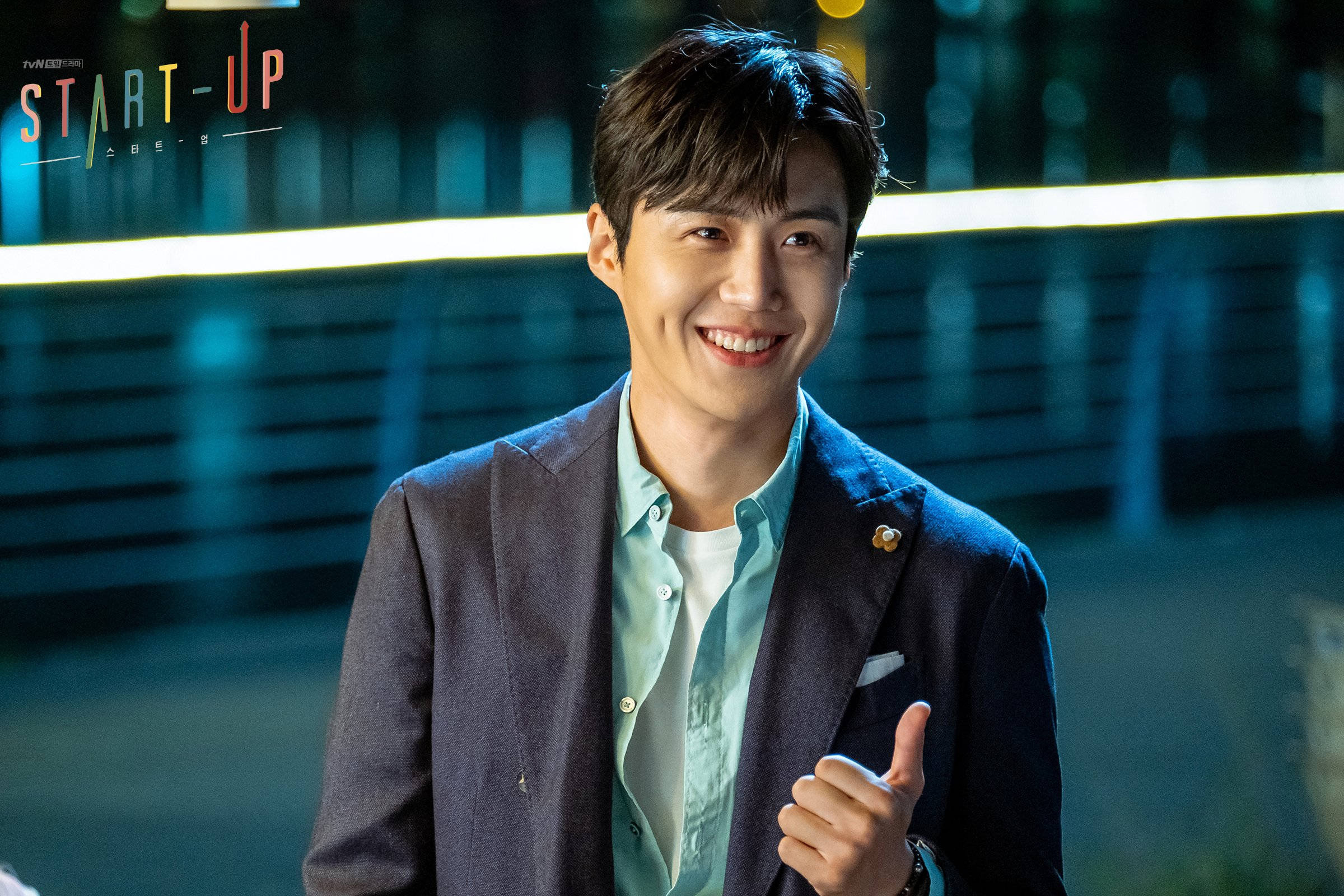 金宣虎《Start Up》「好男孩」造型 壁紙 — 一個微笑的男人穿著西裝外套，在霓虹燈閃爍的城市夜景背景下豎起大拇指。