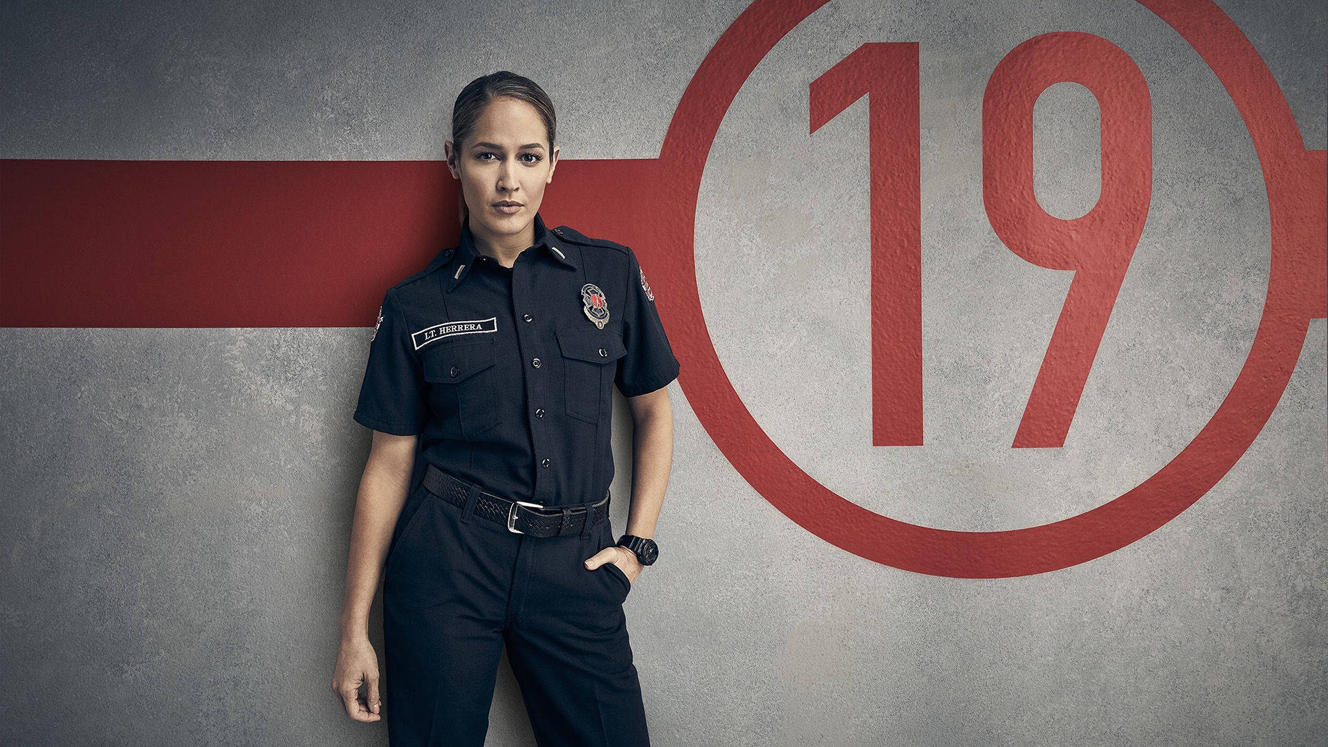 Station 19 中尉安迪·赫雷拉行動照 壁紙 — 一位身穿制服的女消防員自信地站在一面混凝土牆前，牆上漆有一個大大的紅色數字19。