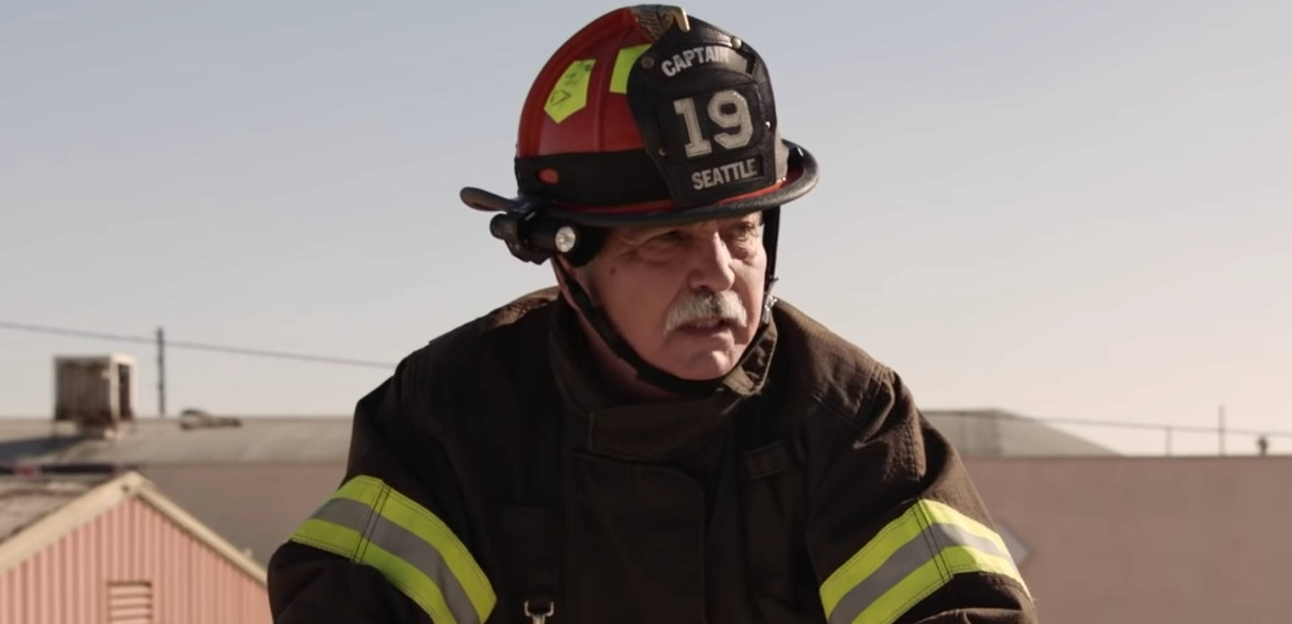 《Station 19》普里特·赫雷拉晉升行動畫面 壁紙 — 一位經驗豐富的西雅圖消防隊長穿著裝備，在晴朗的天空和屋頂背景下專注地向前看。