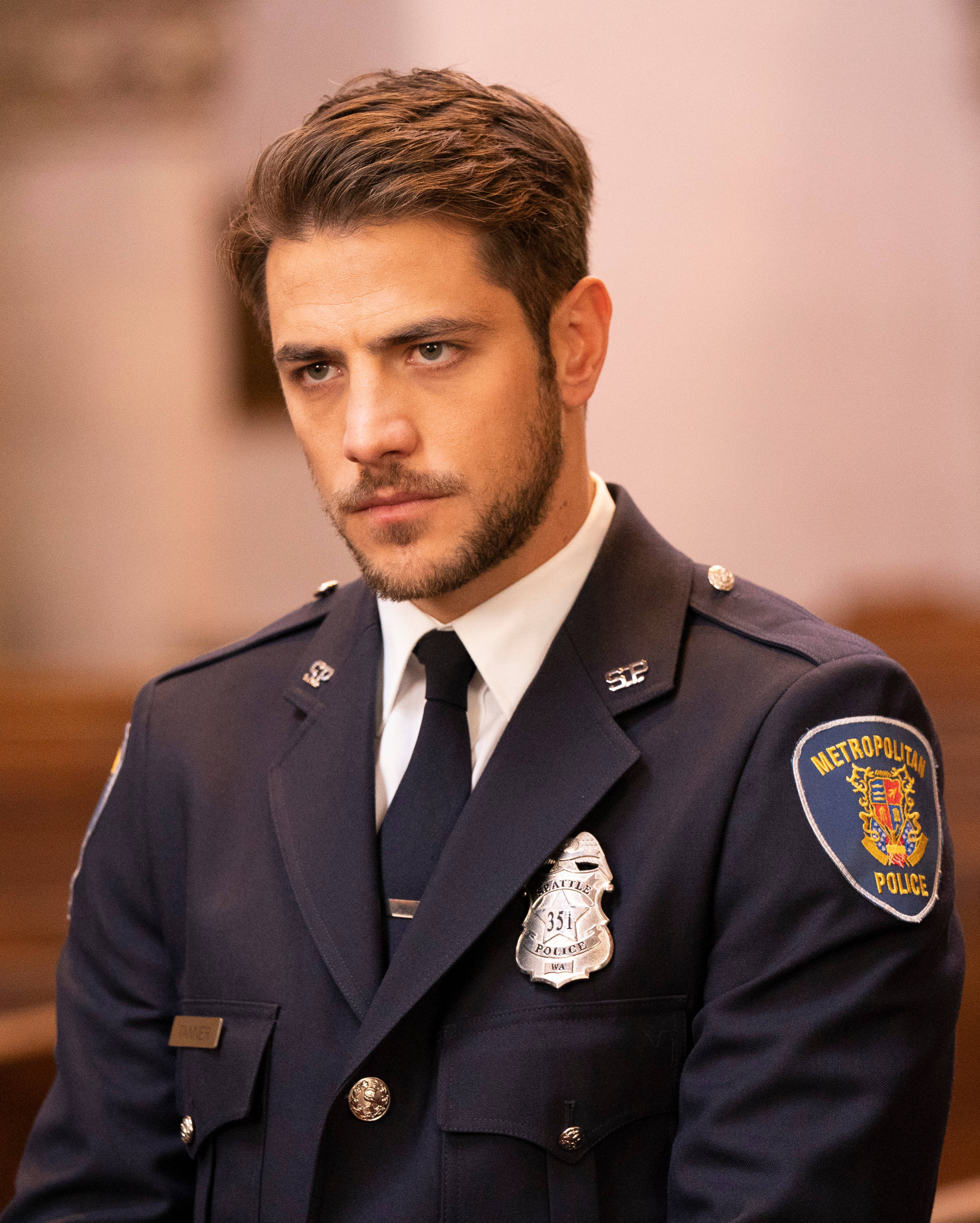 《Station 19》演員亞歷山卓·弗雷薩（飾演瑞安·坦納） 壁紙 — 一名嚴肅的警察穿著深藍色制服，帶有徽章和臂章，站在柔和燈光的室內環境中。