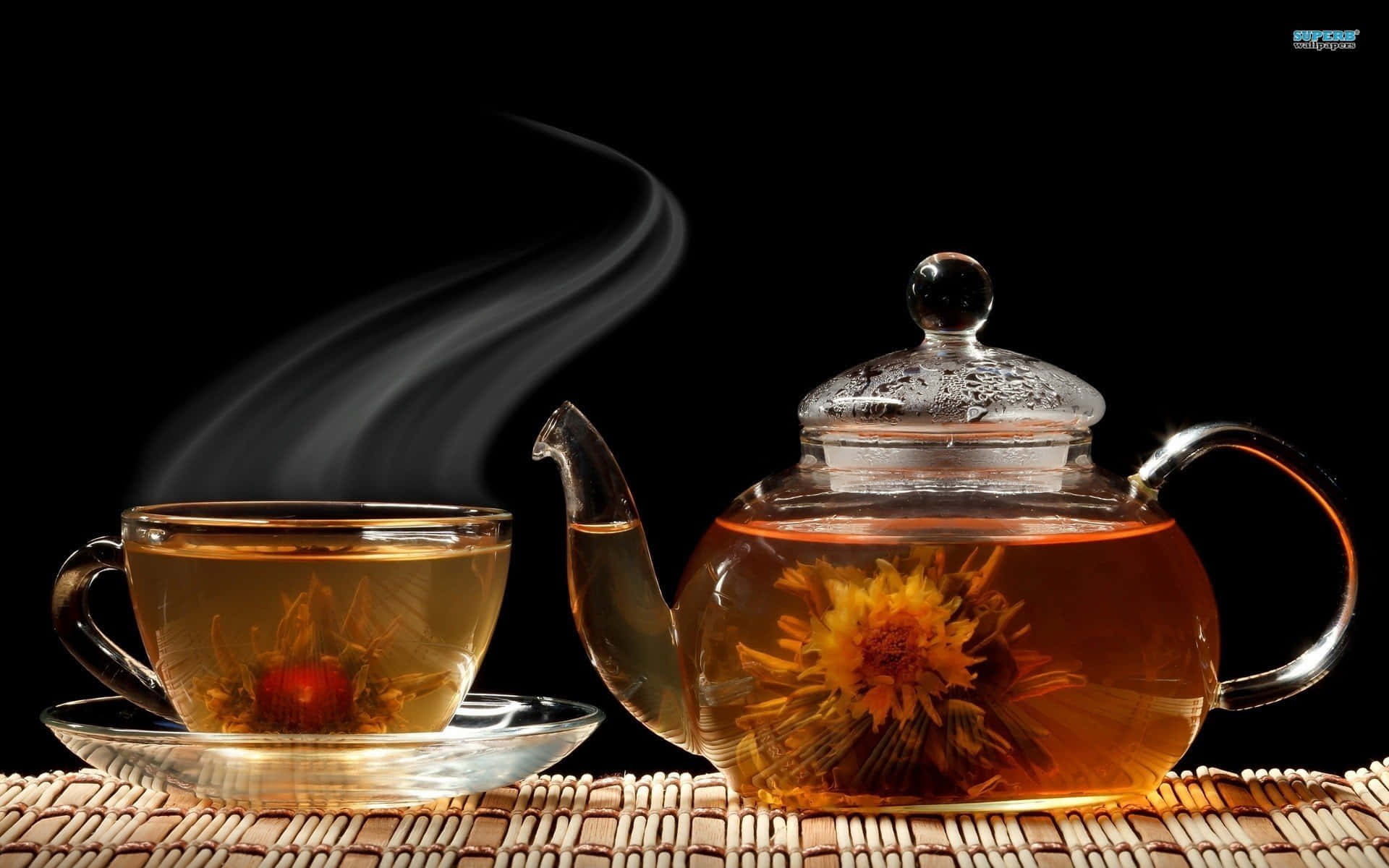 玻璃茶壺和杯子，裝有冒煙的花草茶和盛開的花朵，放在編織墊上。