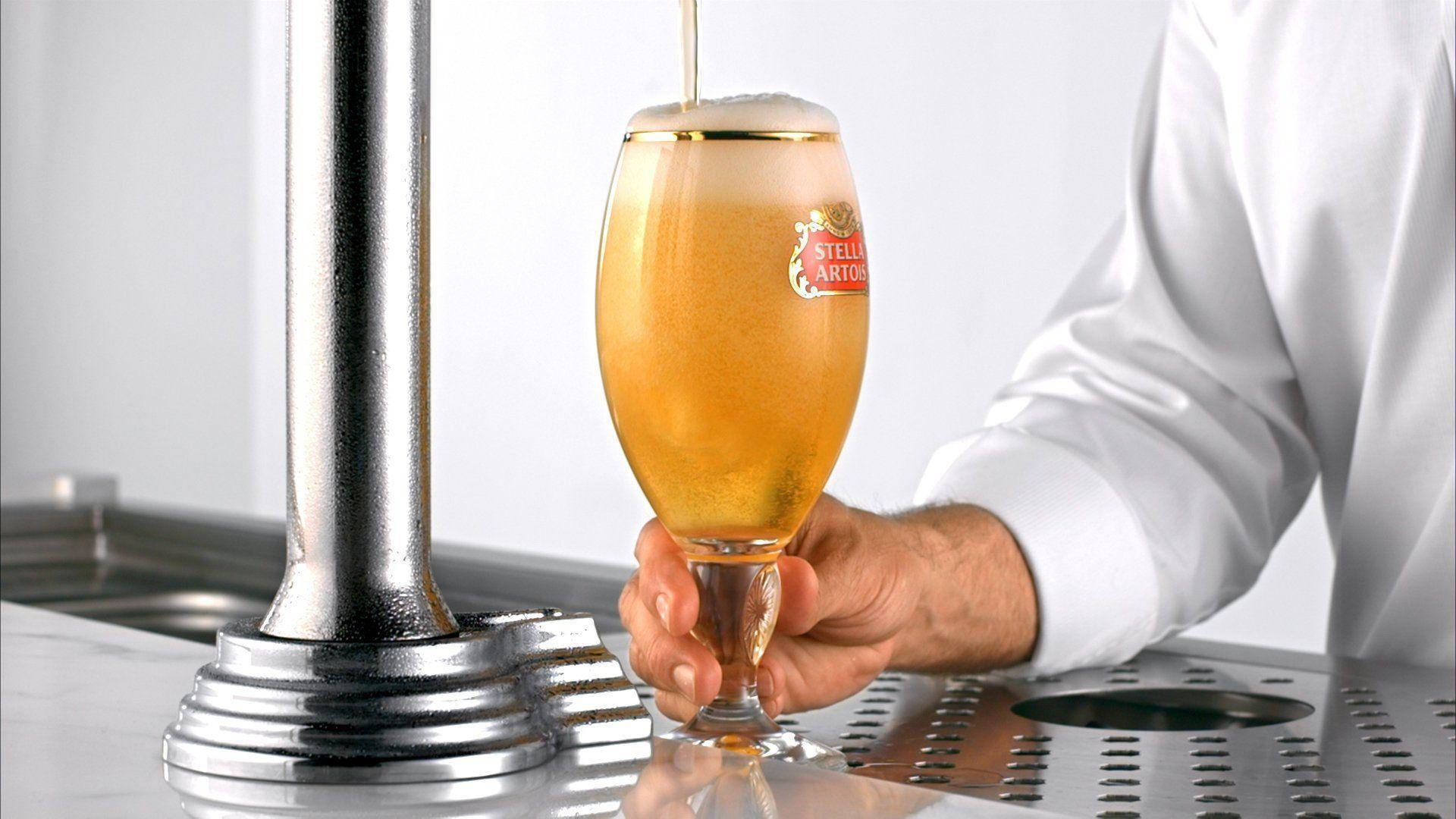 滿杯 Stella Artois 啤酒 壁紙 — 調酒師將金黃色的 Stella Artois 啤酒倒入品牌玻璃杯中，帶有綿密泡沫，背景是乾淨的金屬吧檯。