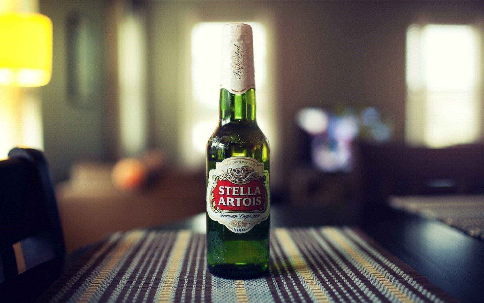 以獲獎殊榮的 Stella Artois 拉格啤酒慶祝品質 壁紙 — 一個綠色 Stella Artois 啤酒瓶放在條紋桌布中央，處於溫暖照明、柔和模糊的客廳中。
