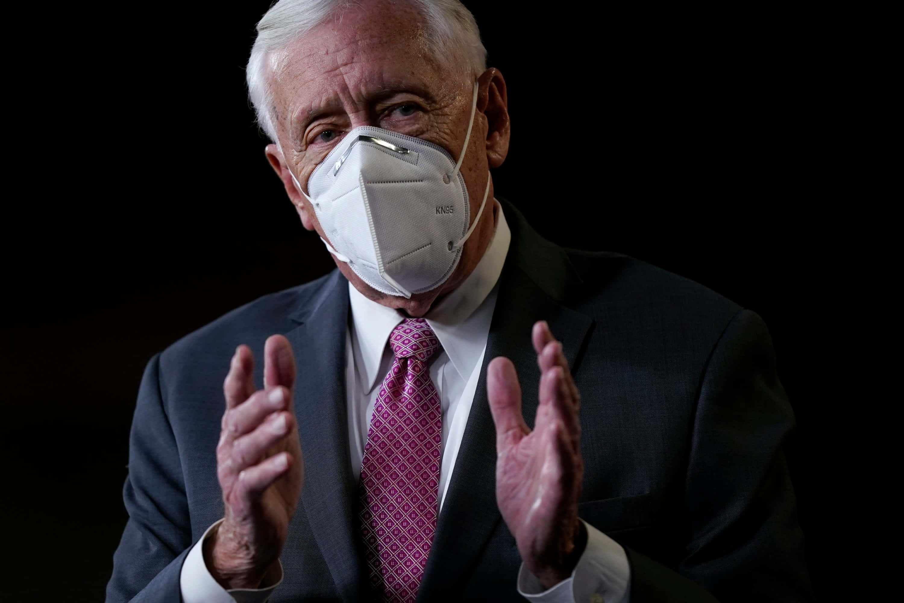 Steny Hoyer 戴口罩做手勢 壁紙 — 一名身穿西裝和粉紅色領帶的男子在黑色背景前戴著白色口罩做手勢。