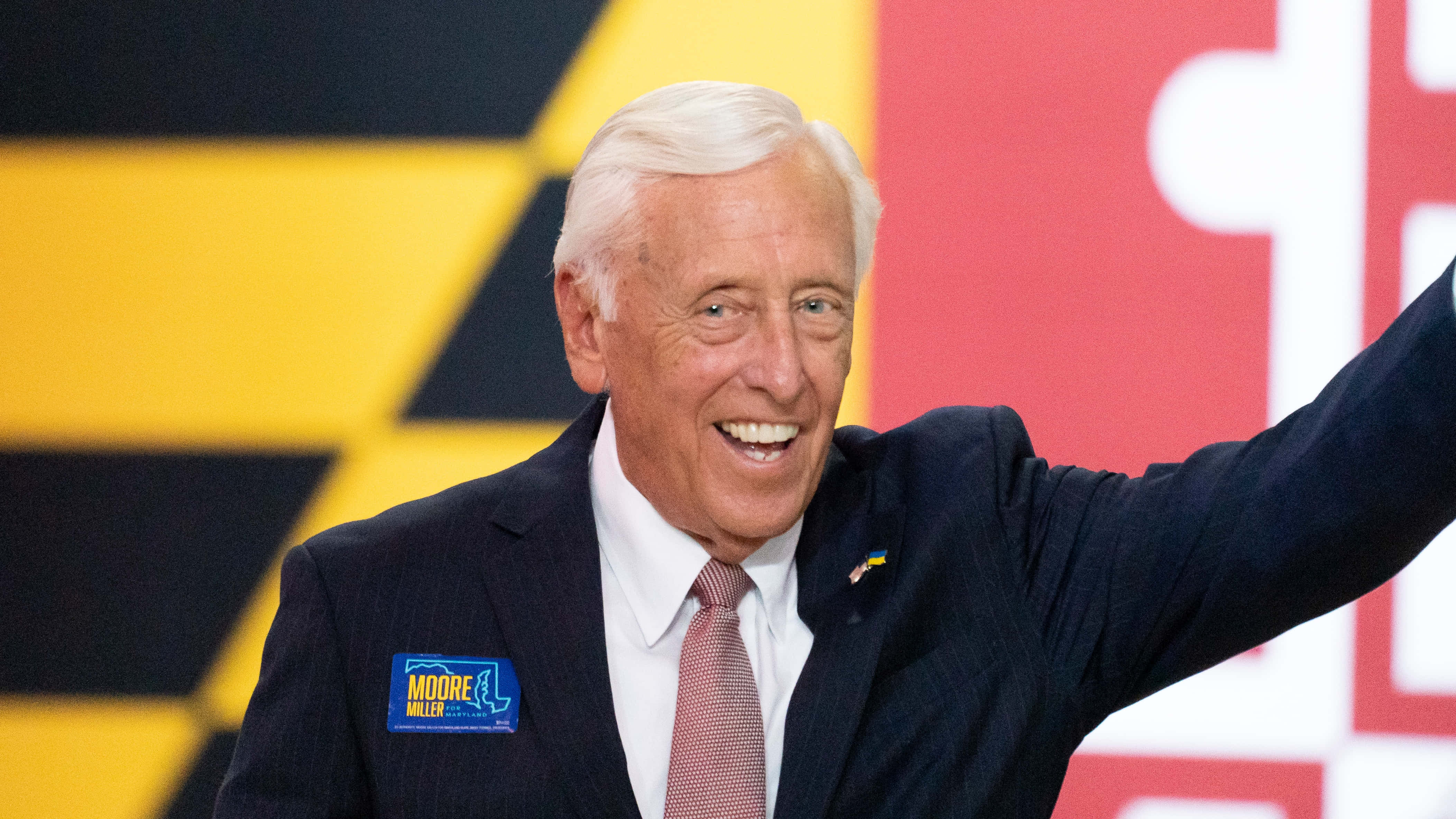 Steny Hoyer 微笑向觀眾揮手 壁紙 — 一位白髮老人身穿深色西裝，在鮮明的黃、黑、紅背景前微笑揮手。