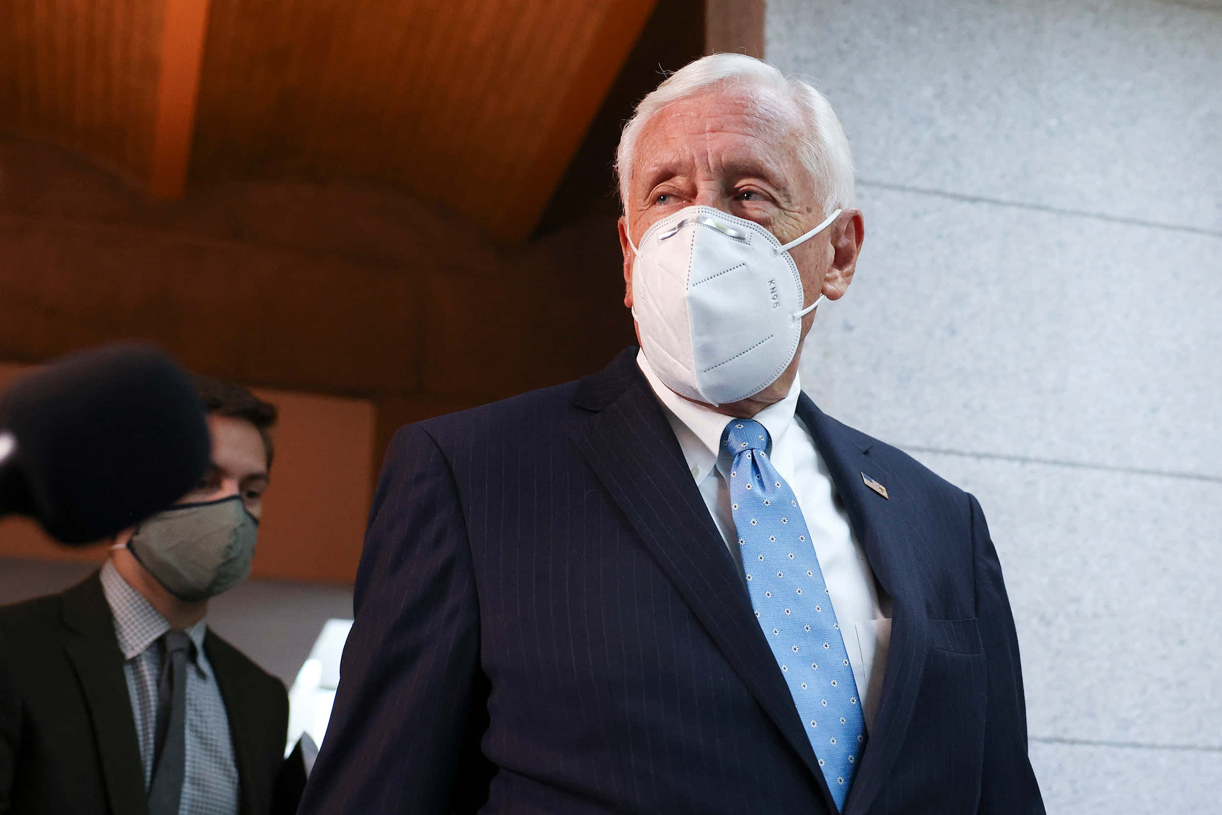 Steny Hoyer 戴口罩行走 壁紙 — 一位身穿深色西裝和淺藍色領帶的年長男性戴著白色口罩，走過一個麥克風和另一個戴口罩的人。