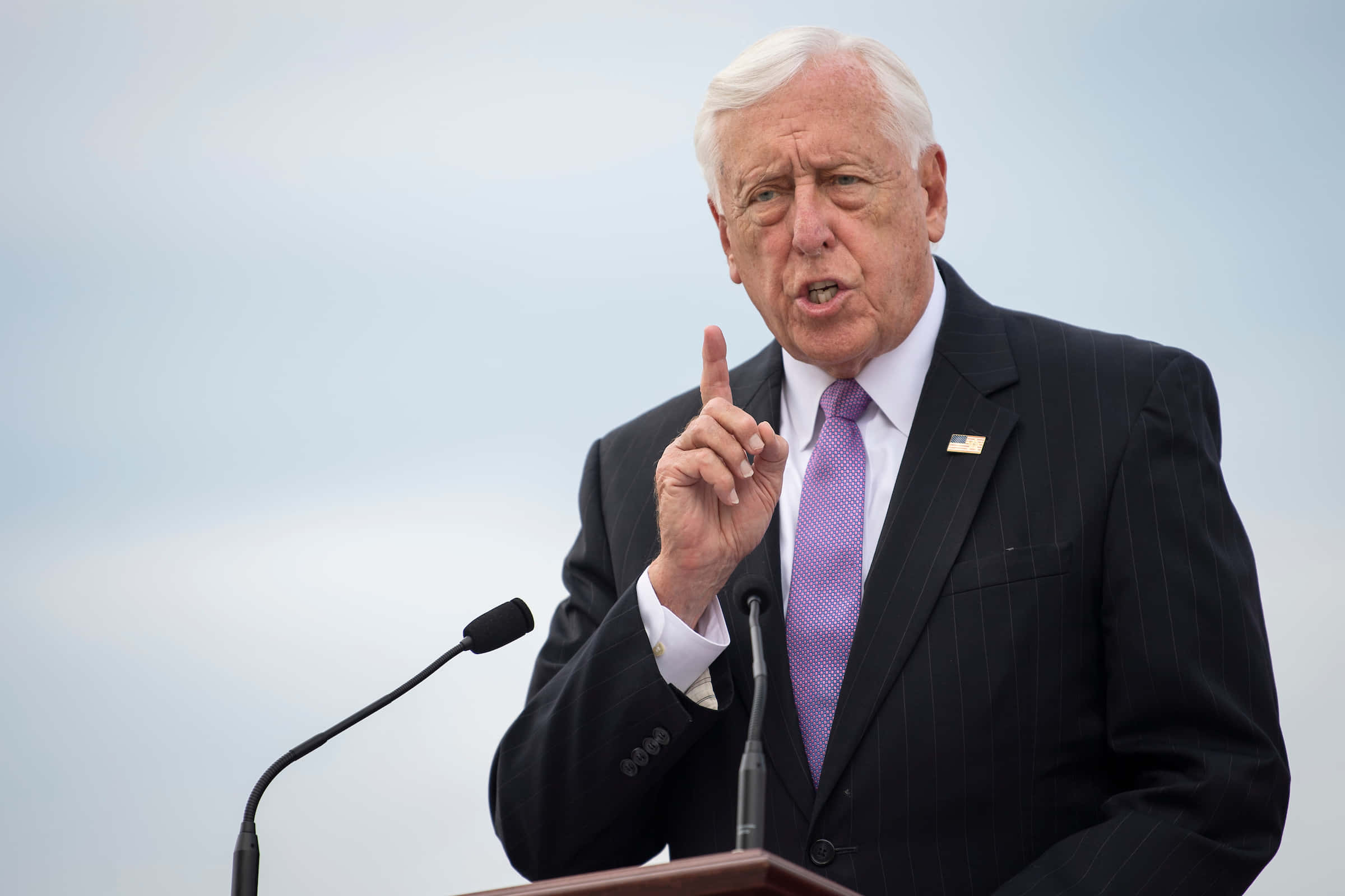 Steny Hoyer 做出強烈論辯姿勢 壁紙 — 一位年長男士穿著深色細條紋西裝和紫色領帶，在蒼白的天空下於戶外講台上激動地比劃著手勢發言。