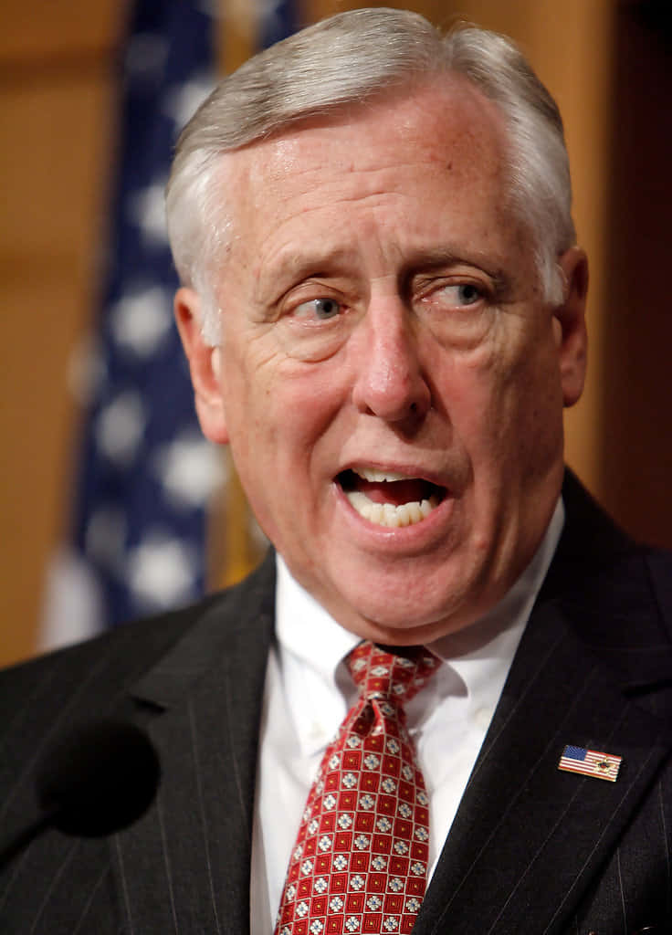 Steny Hoyer 偷笑並說話 壁紙 — 近距離拍攝一名身穿西裝的男子，熱情地演講，佩戴美國國旗別針，背景有模糊的旗幟。