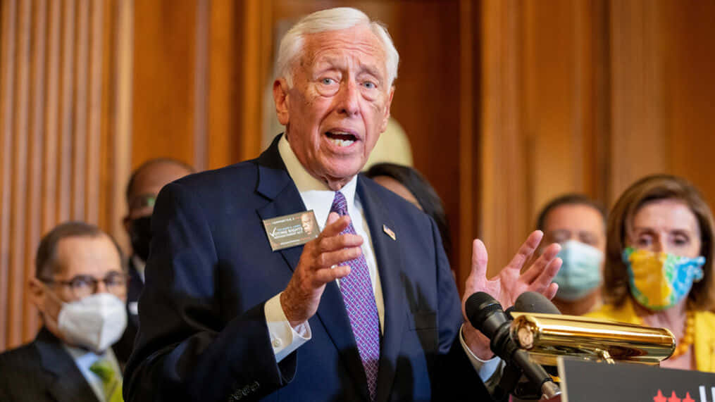 Steny Hoyer 在熱情的觀眾前發表講話 壁紙 — 一位身穿深色西裝和圖案領帶的年長男性在講台上激情演講，身後有戴著口罩的人們在聆聽。