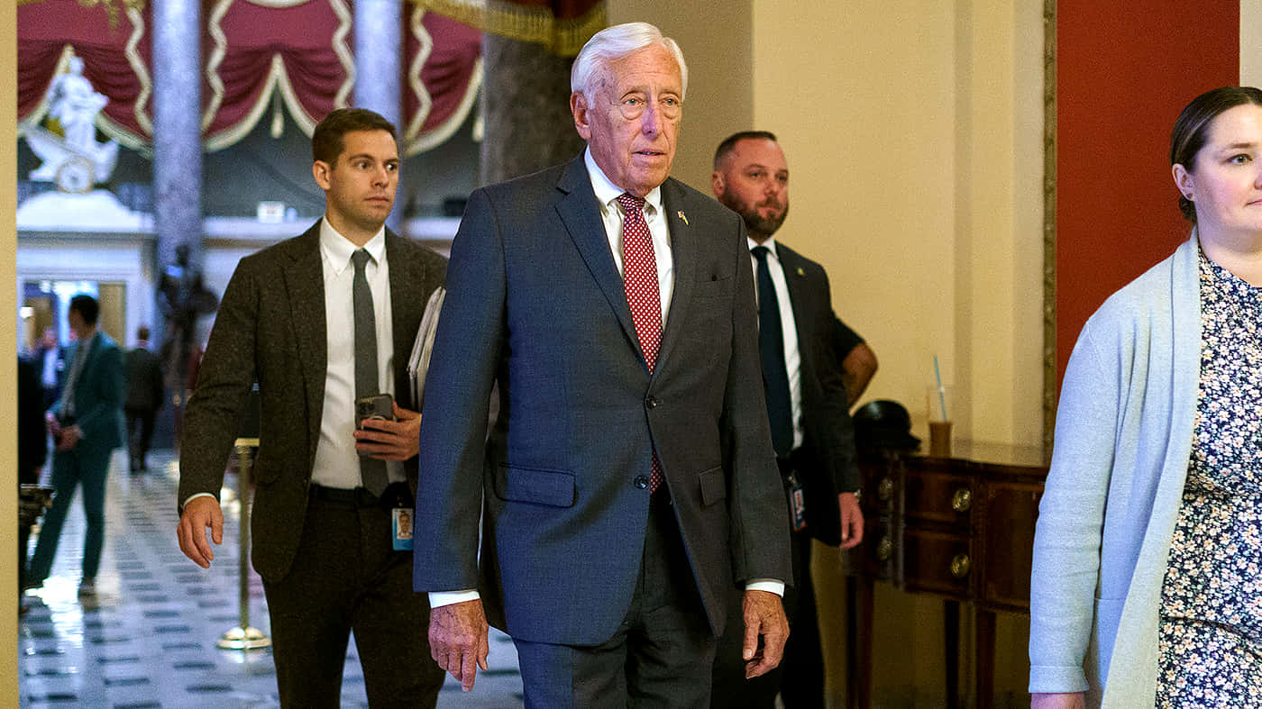 Steny Hoyer 在走廊中行走 壁紙 — 一位年長男士身穿深色西裝和紅色領帶，在保安和職員的陪同下走過宏偉的走廊。