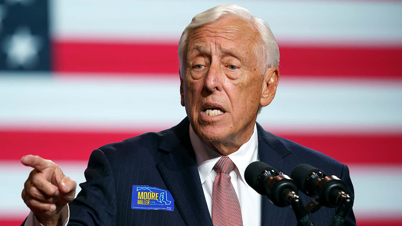 Steny Hoyer 演講時指著前方 壁紙 — 一位身穿西裝的年長男性在模糊的美國國旗背景下，在設有咪高峰的講台上強調地做手勢並發言。