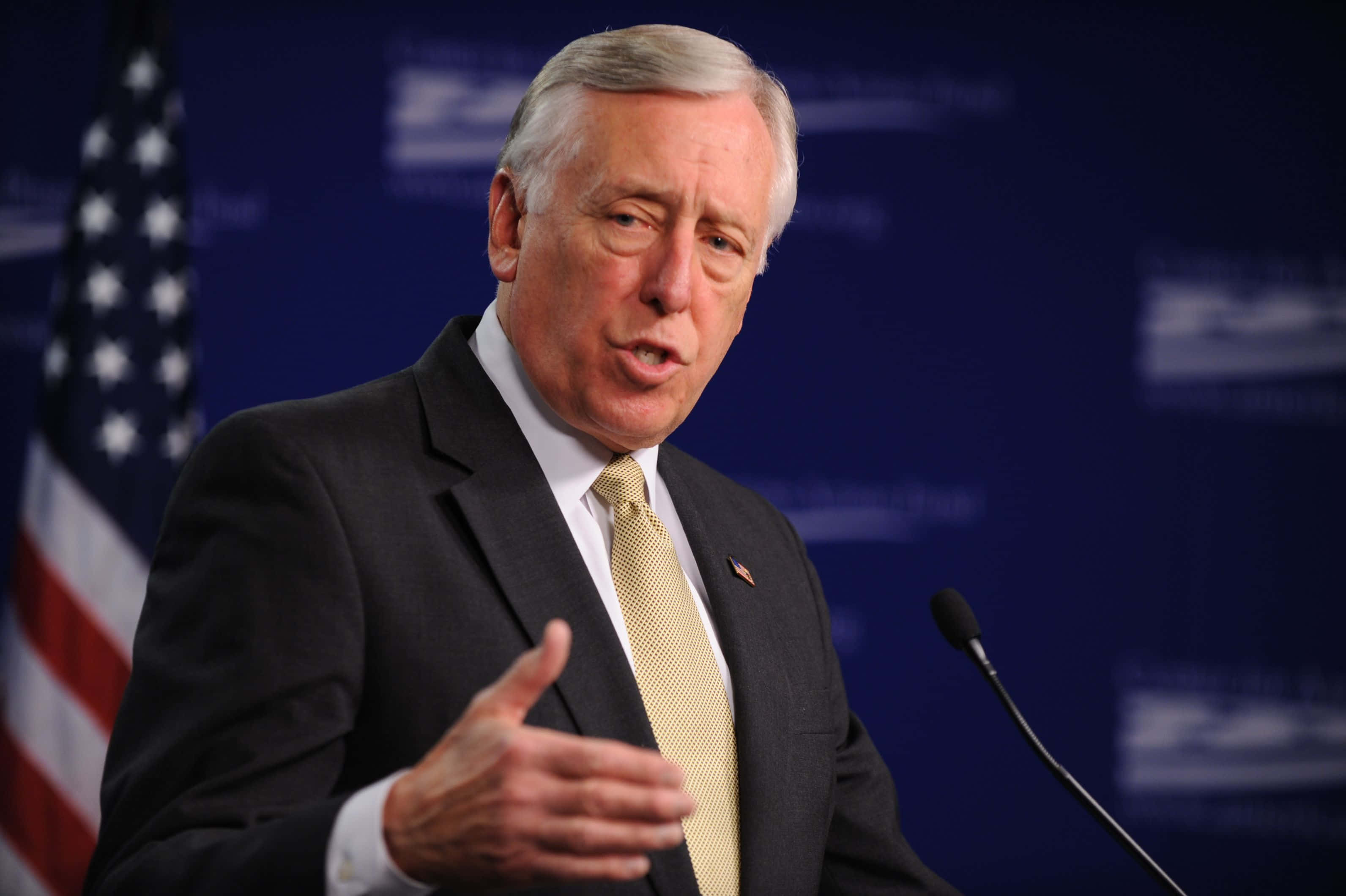Steny Hoyer 演講時語氣流暢 壁紙 — 一名身穿深色西裝和黃色領帶的男子在講台上發言，身後有美國國旗，背景為藍色。