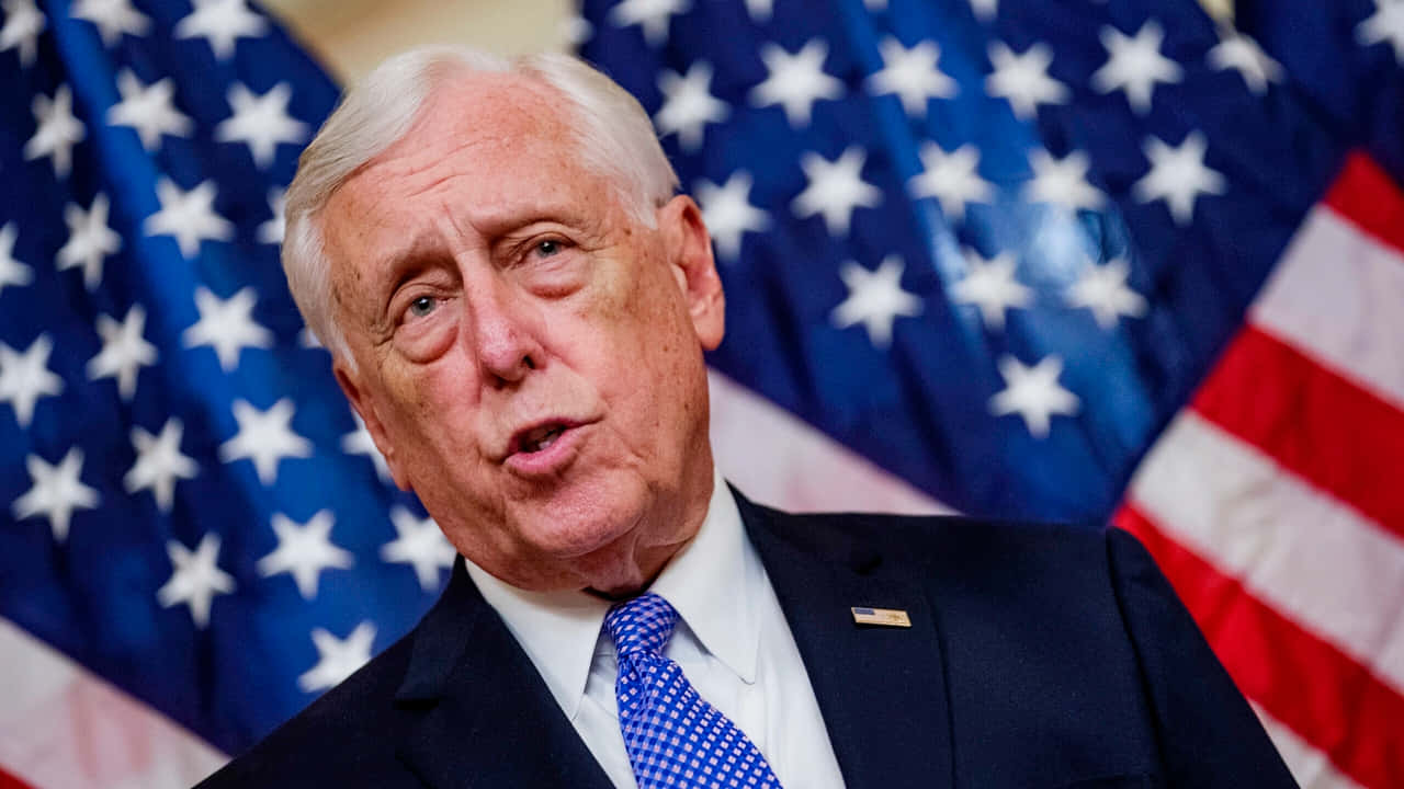 Steny Hoyer 背對美國國旗 壁紙 — 一位身穿西裝和藍色領帶的長者男士在美國國旗背景下說話的特寫。