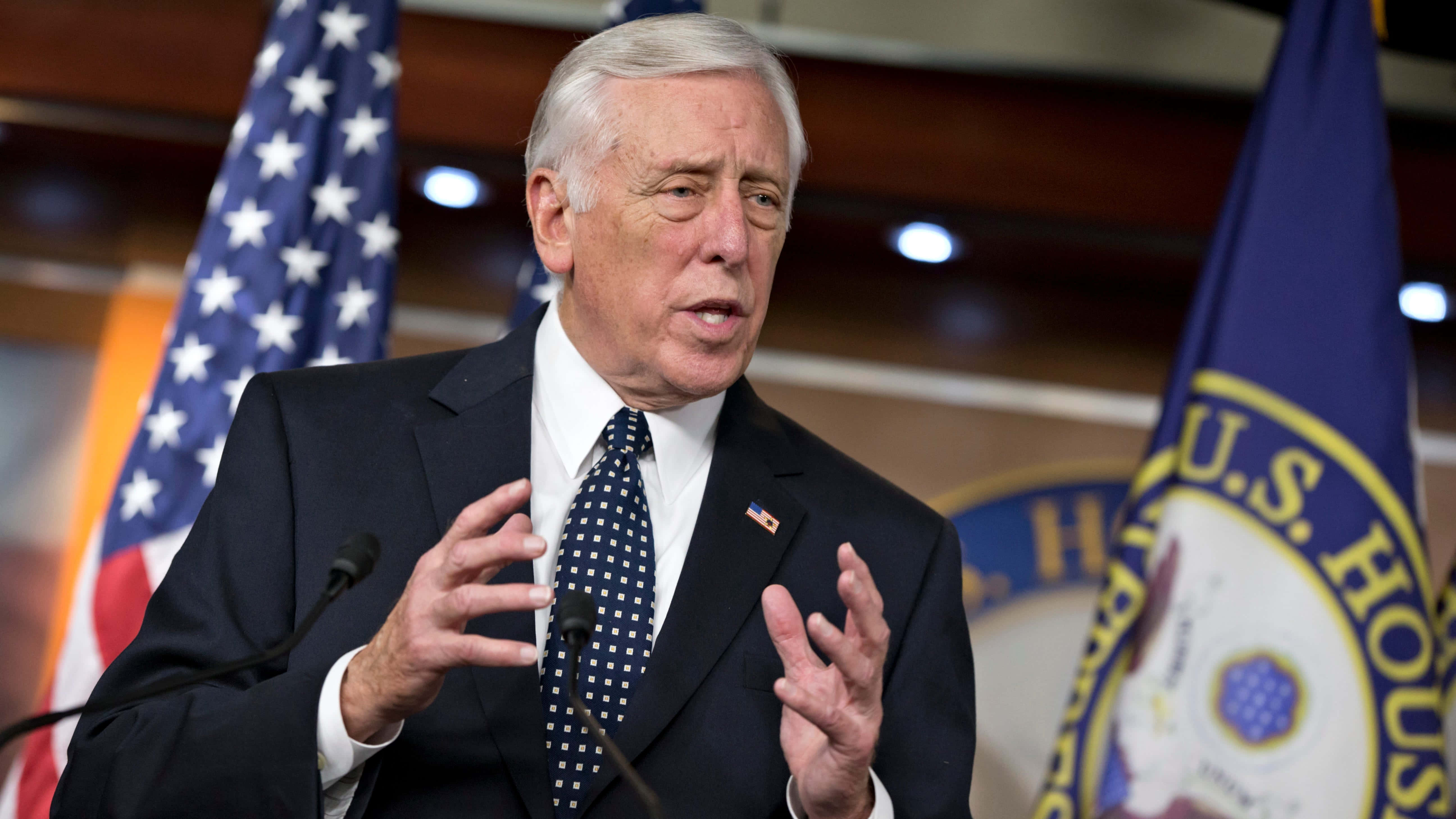 Steny Hoyer 與旗幟對話 壁紙 — 一位年長男士身穿深色西裝和圖案領帶，在兩旁有美國國旗和美國眾議院旗幟的講台上熱情演講。