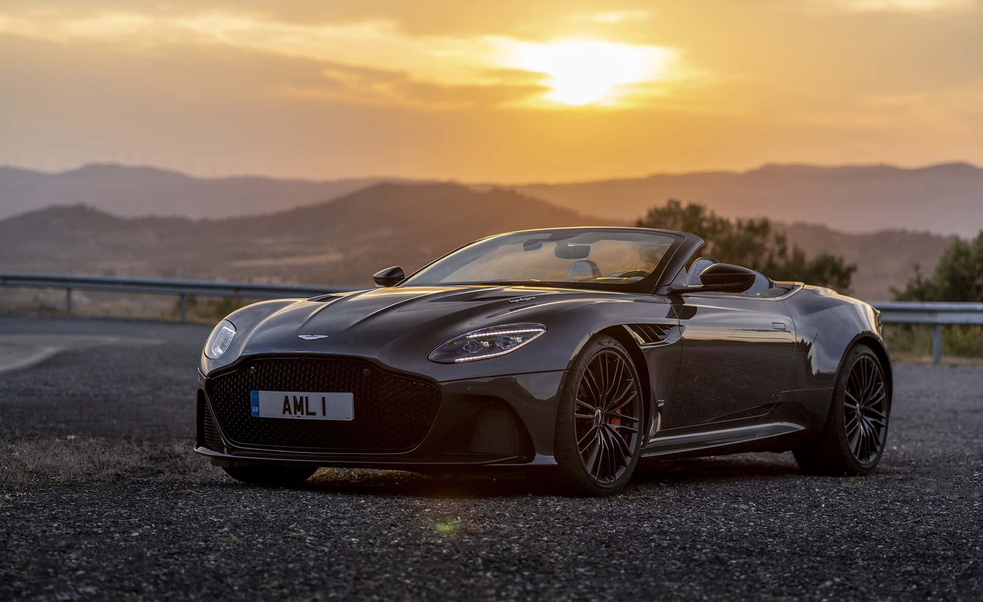 Aston Martin Vantage 桌布 壁紙 — 一架時尚的黑色Aston Martin DB11 Volante開篷車停泊在碎石路上，背景是日落時分的山脈。