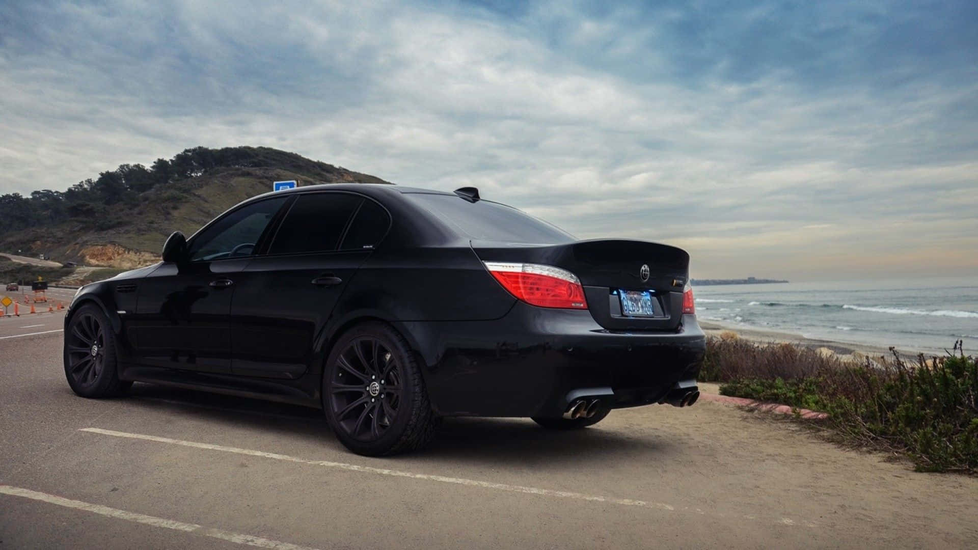 Bmw M5 桌布 壁紙 — 一架時尚的黑色BMW轎車停泊在沿海公路上，海浪在附近拍打，天空多雲。