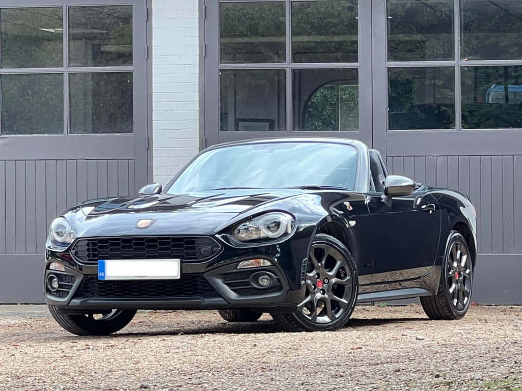 Fiat 124 Spider 桌布 壁紙 — 一架時尚黑色快意124 Spider開篷車停泊在現代灰色車庫前的碎石地上，車庫有大型窗戶。
