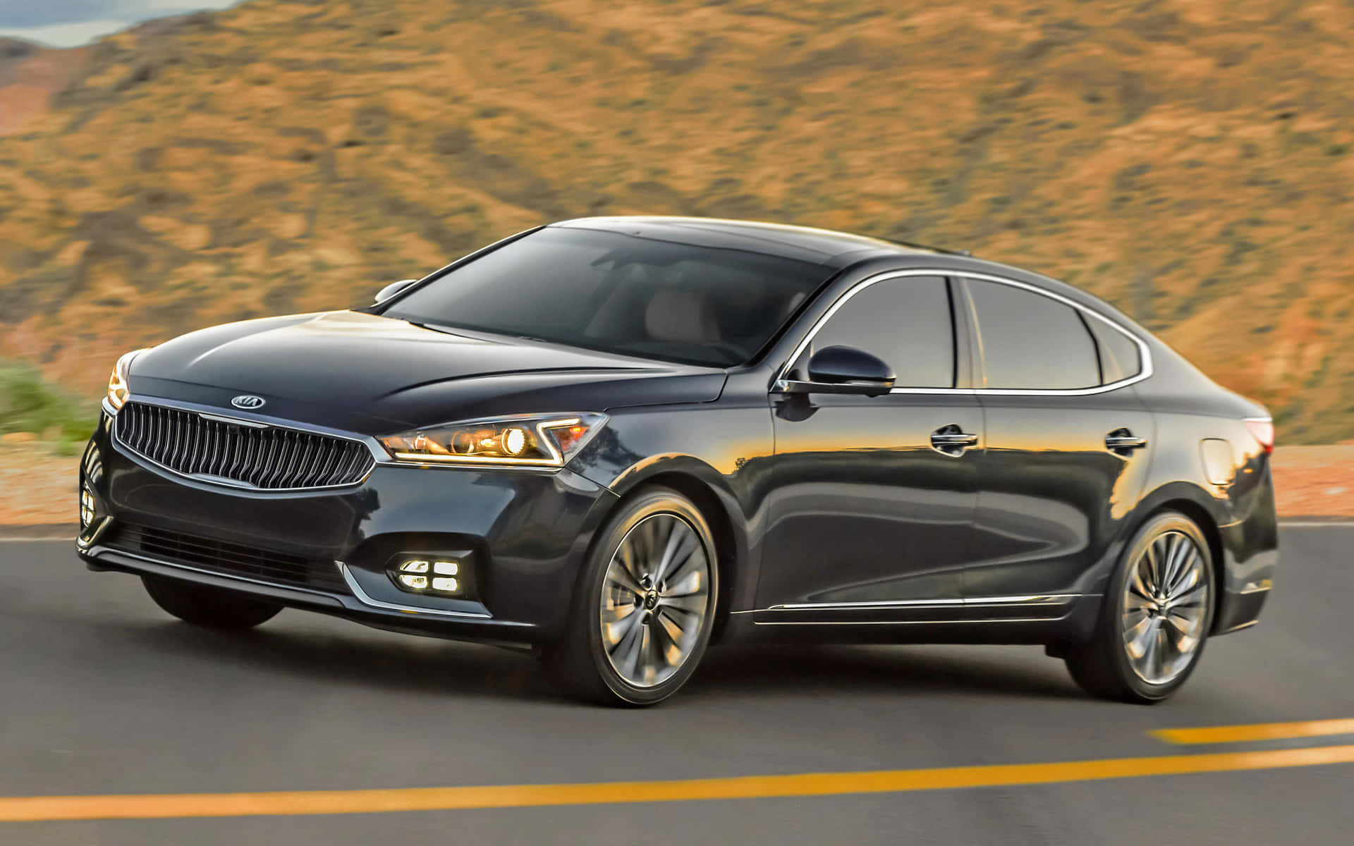 Kia Cadenza 桌布 壁紙 — 一輛時尚的黑色Kia轎車在沙漠公路上行駛，背景是金色的山丘。